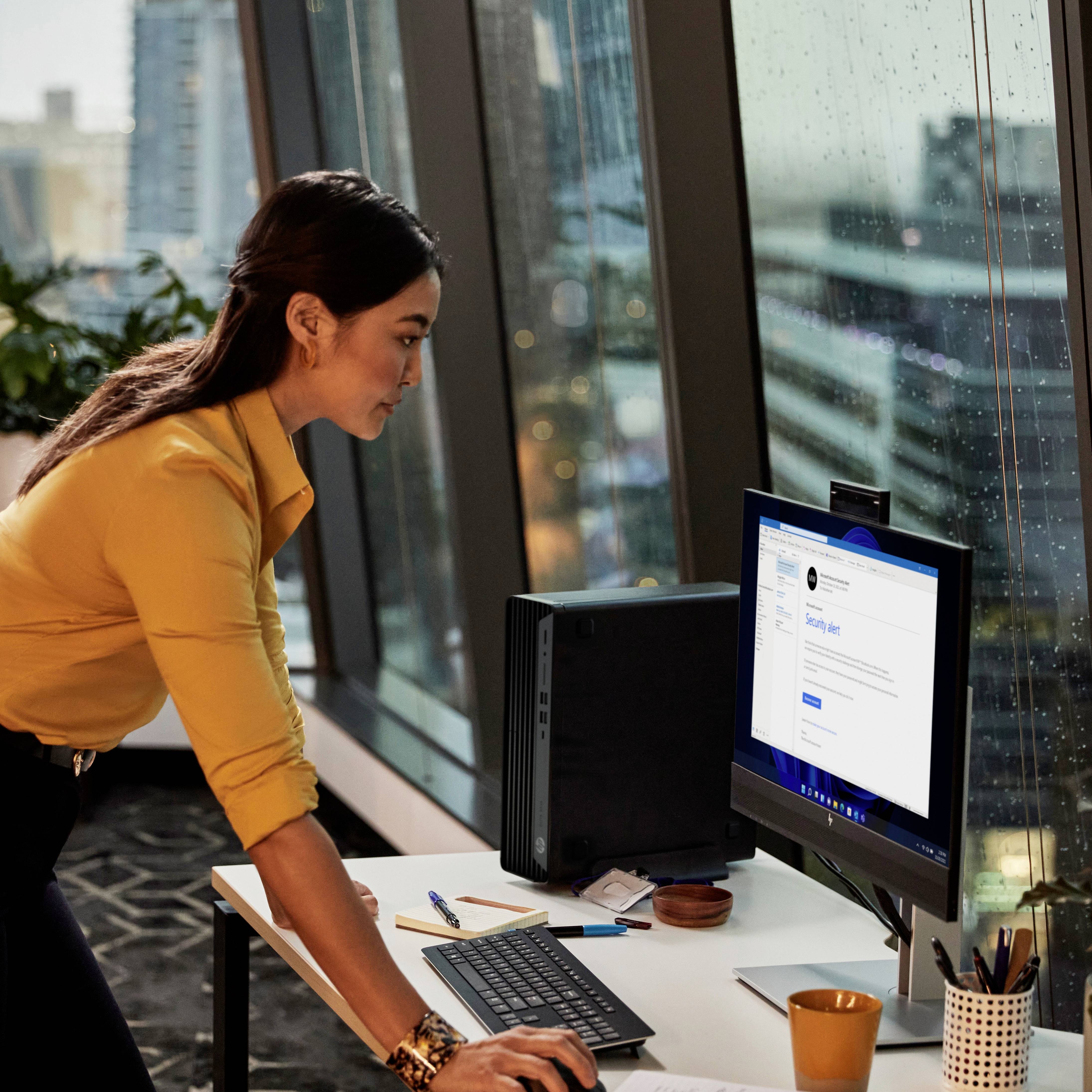 Eine Frau in gelber Bluse arbeitet an einem Computer mit einem modernen Büro im Hintergrund. Sie betrachtet konzentriert den Bildschirm.