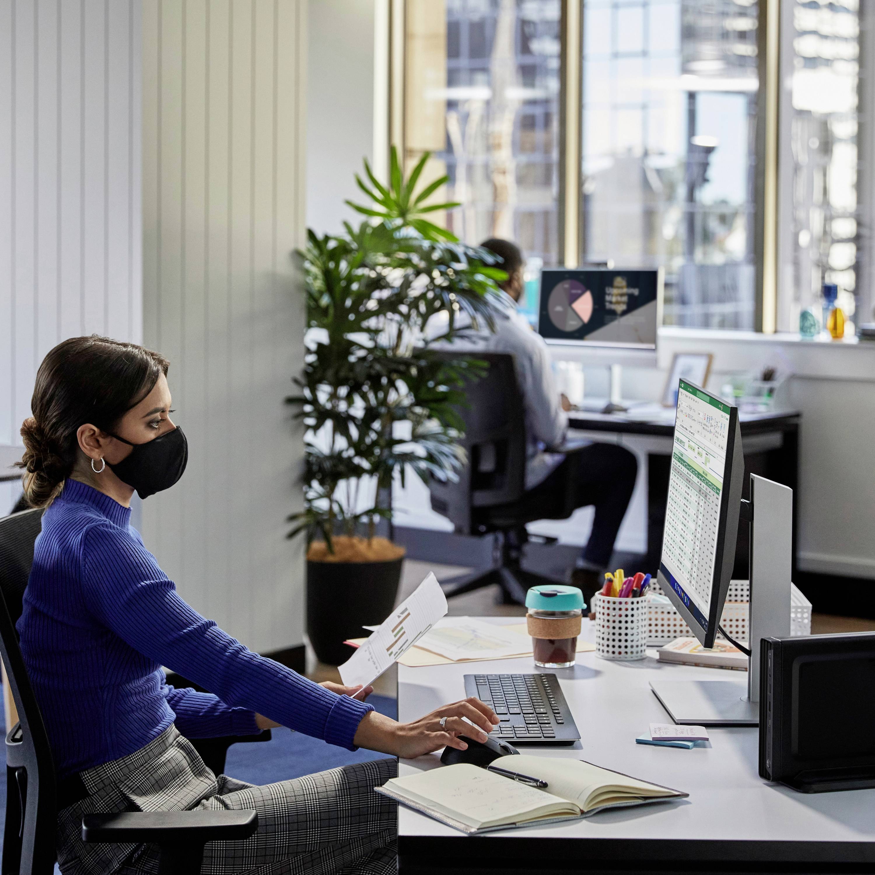 Eine Frau mit Maske arbeitet an einem Schreibtisch im Büro. Sie hält Dokumente und blickt auf einen Computerbildschirm mit Tabellenkalkulation.