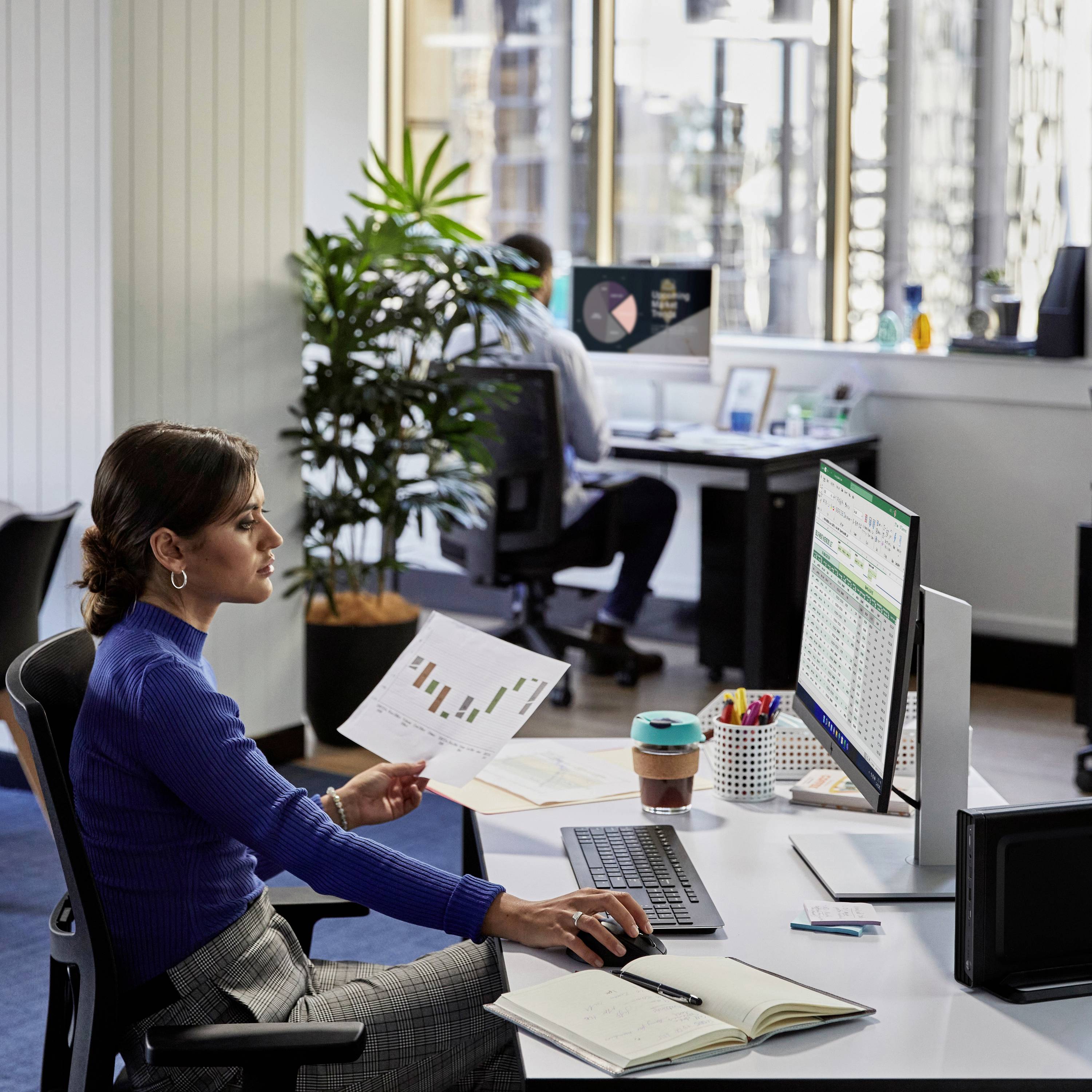 Eine Frau sitzt an einem Bürotisch vor einem Computerbildschirm mit Tabellenkalkulationen. Sie hält ein Diagrammblatt, während ein Kollege im Hintergrund am Laptop arbeitet.