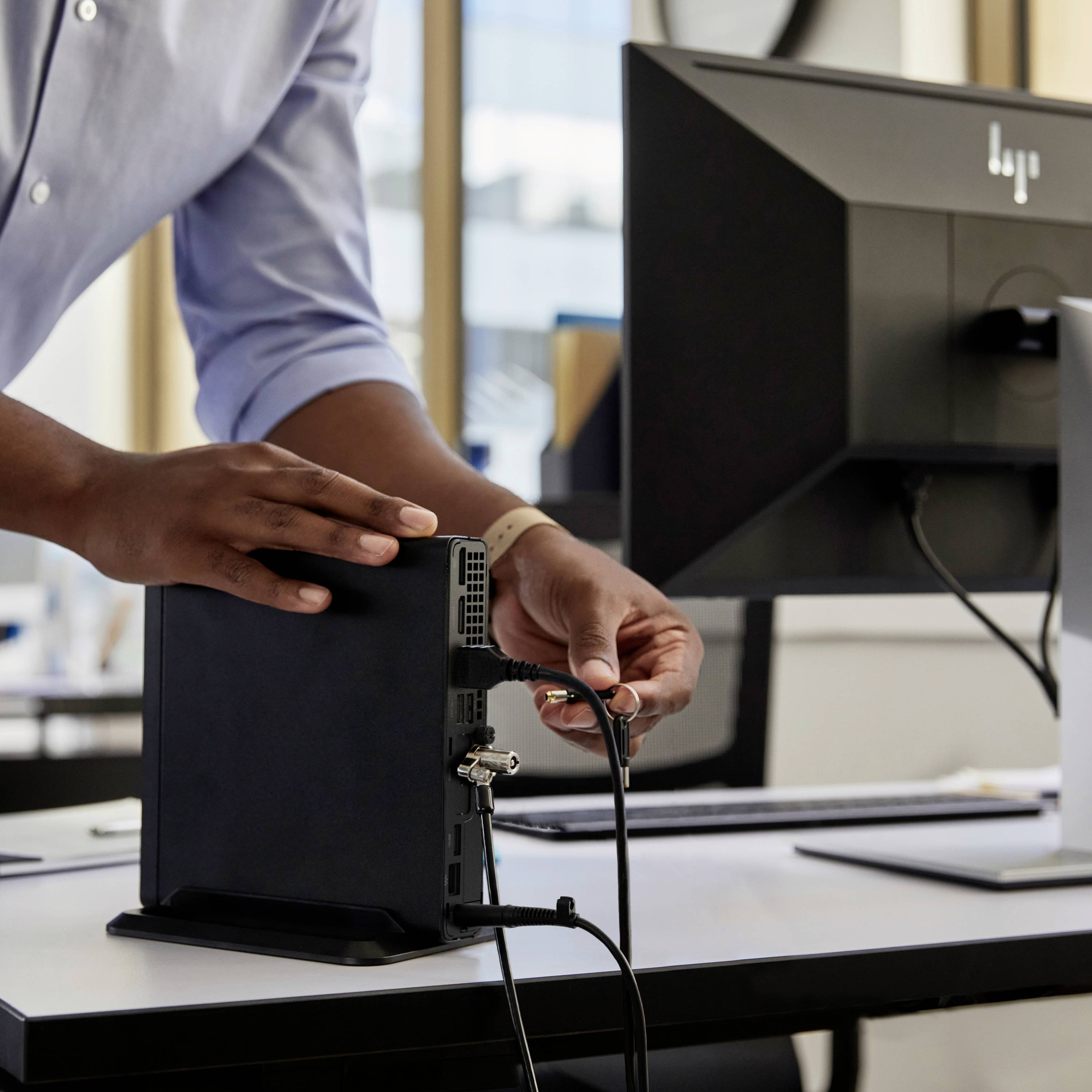 Person schließt Kabel an kleinen Computer an, auf modernem Schreibtisch; im Hintergrund Monitor. Szene in moderner Büroatmosphäre.