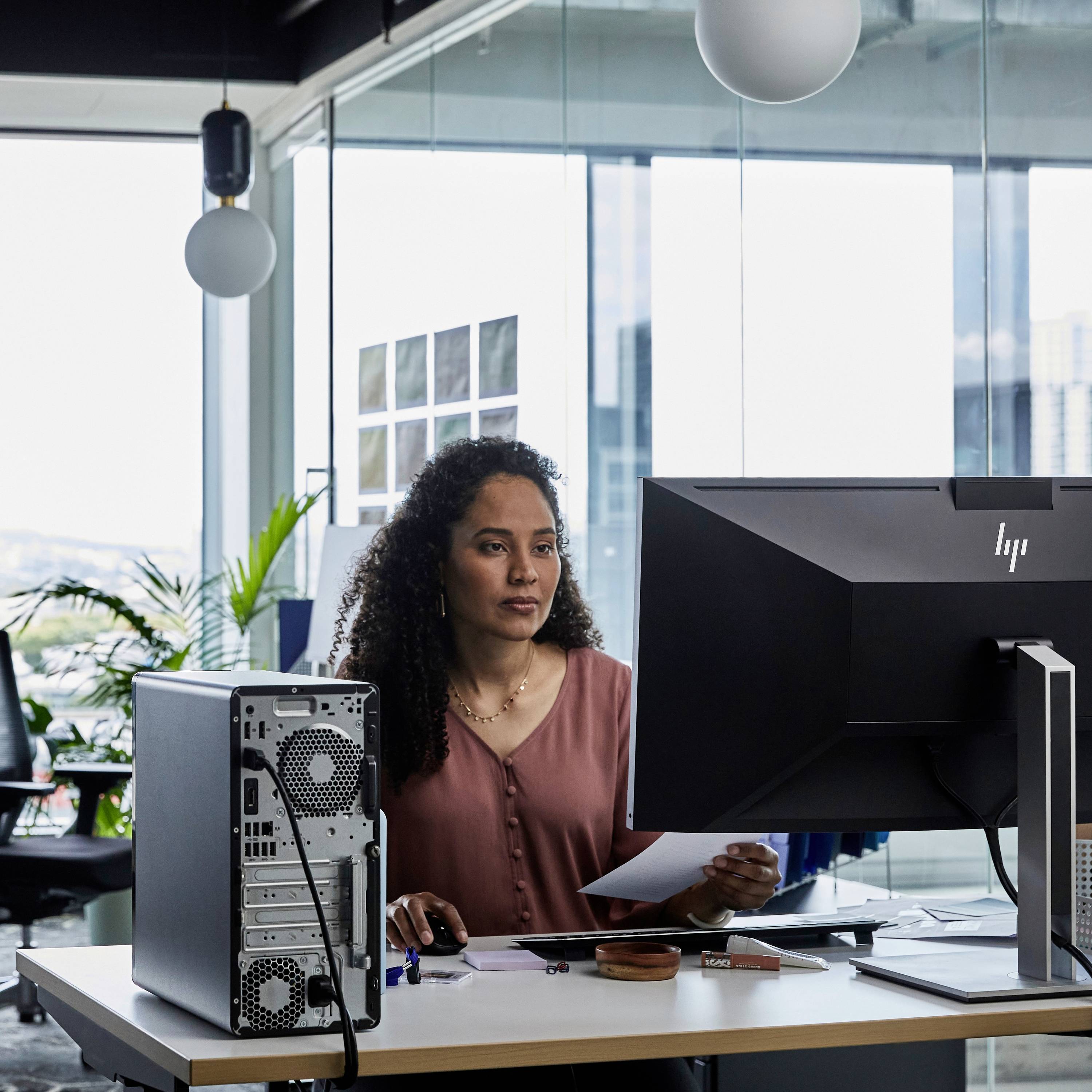 Eine Frau sitzt an einem Schreibtisch mit Computern in einem modernen Büro. Sie blickt auf einen Bildschirm und hält einen Stift und Papier.