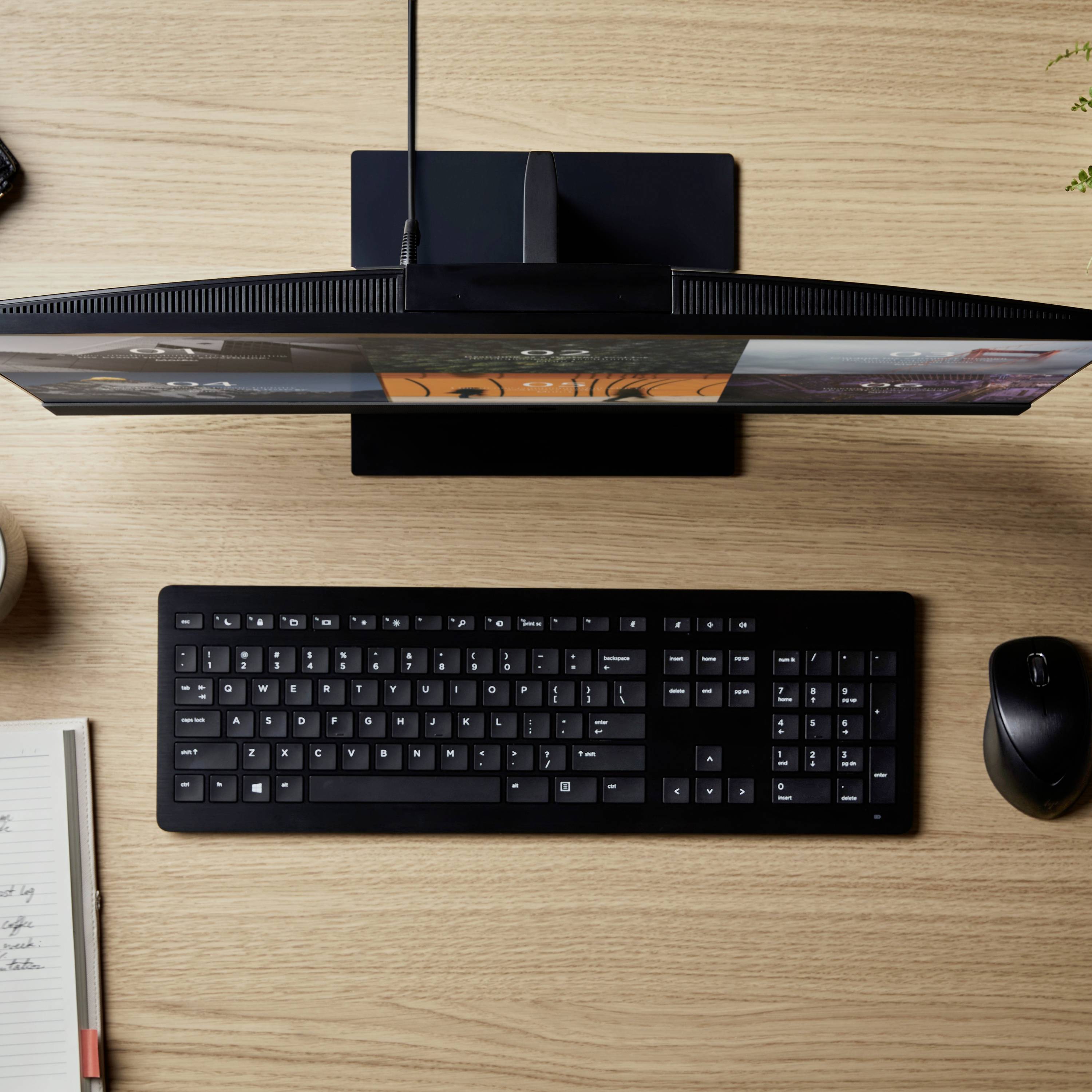 Ein Computerbildschirm von oben betrachtet, mit Tastatur und Maus daneben auf einem Holztisch. Daneben ein Notizblock und eine Tasse.