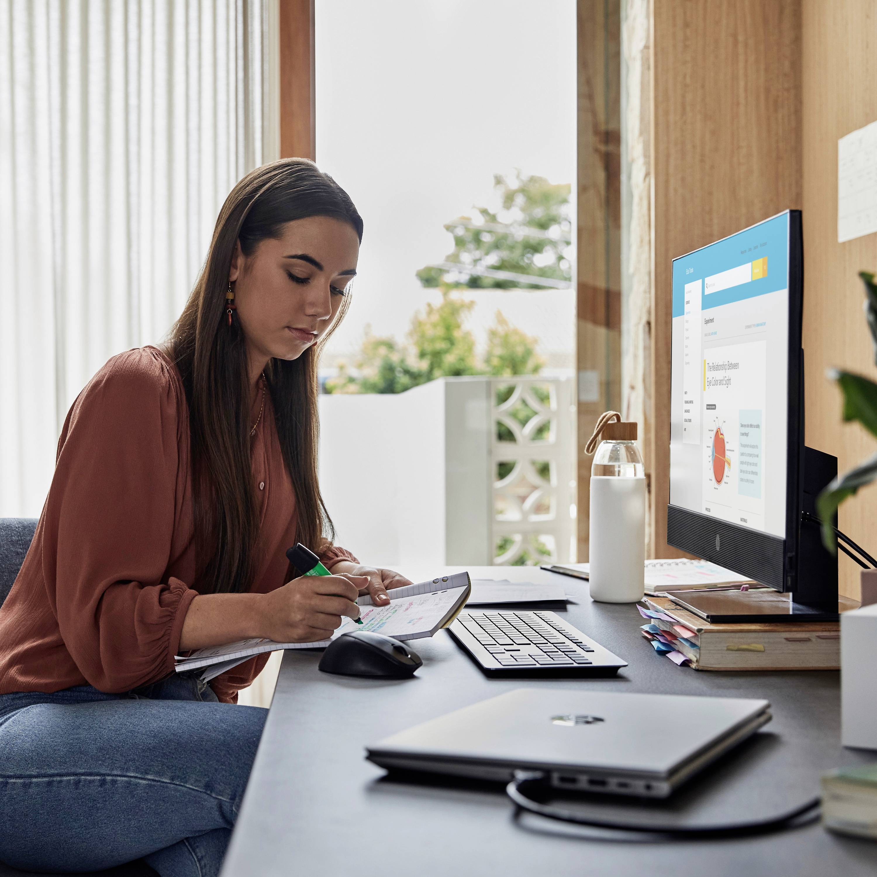 Eine Frau arbeitet konzentriert an einem Schreibtisch mit Laptop und Tablet, umgeben von Schreibmaterialien. Sonnenlicht scheint durch ein Fenster.