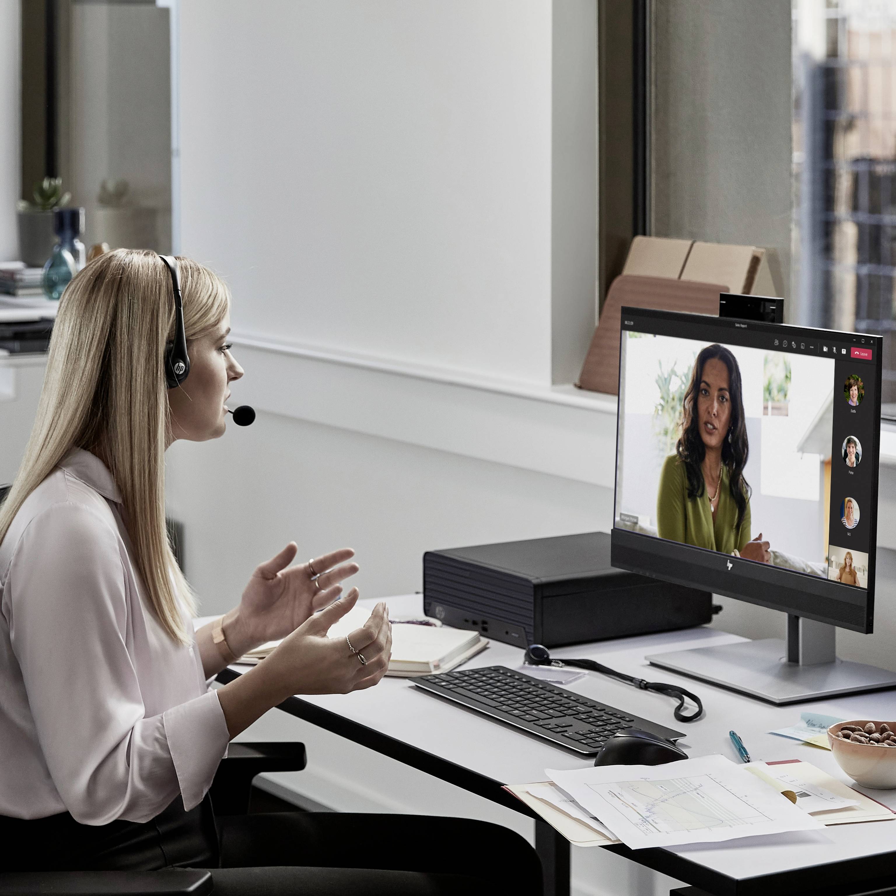 Eine Frau mit Headset führt ein Videomeeting. Auf dem Bildschirm sind drei Teilnehmer sichtbar. Sie sitzt an einem aufgeräumten Schreibtisch.