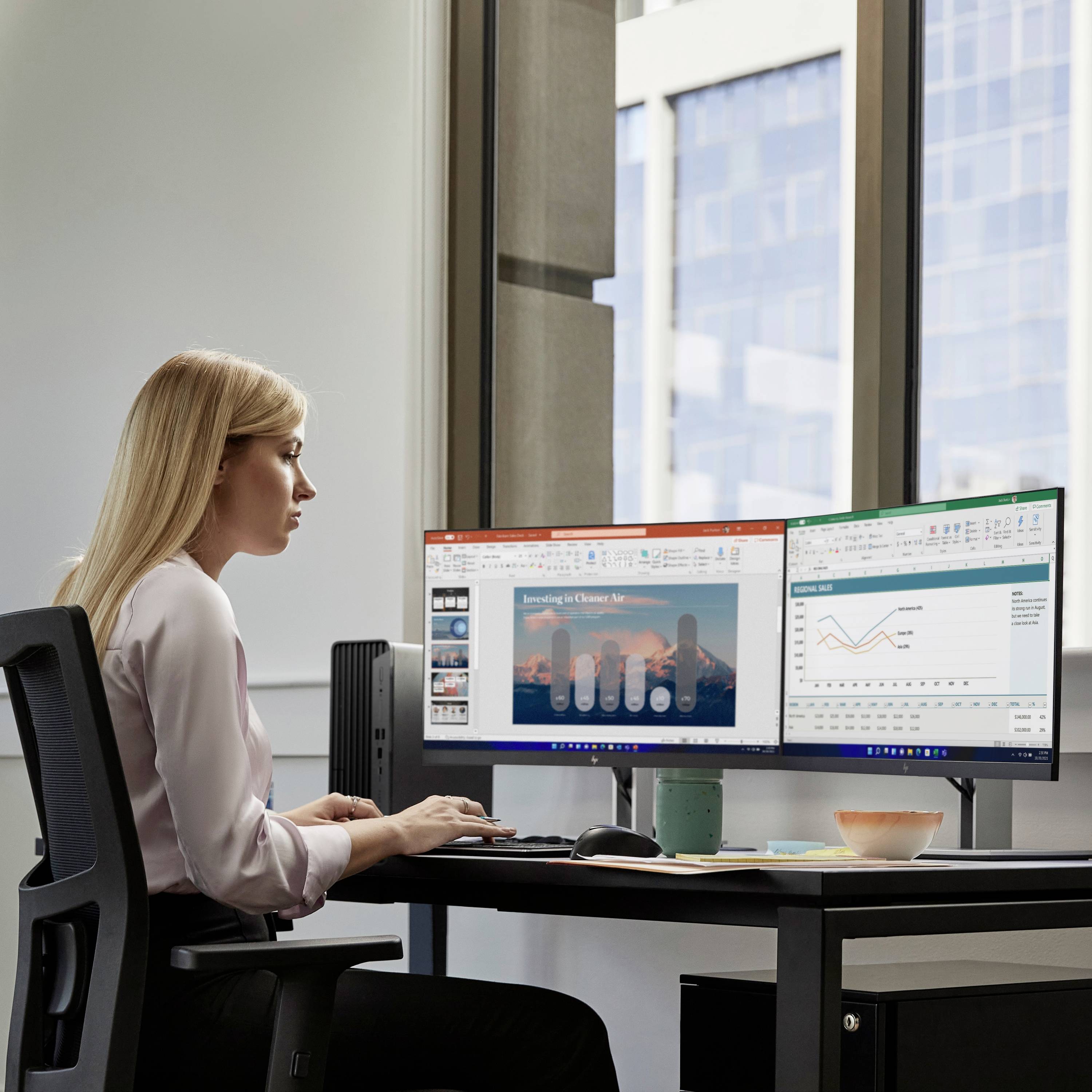Eine Person sitzt an einem Schreibtisch und arbeitet an zwei Monitoren. Auf den Bildschirmen sind Diagramme und Text zu sehen.