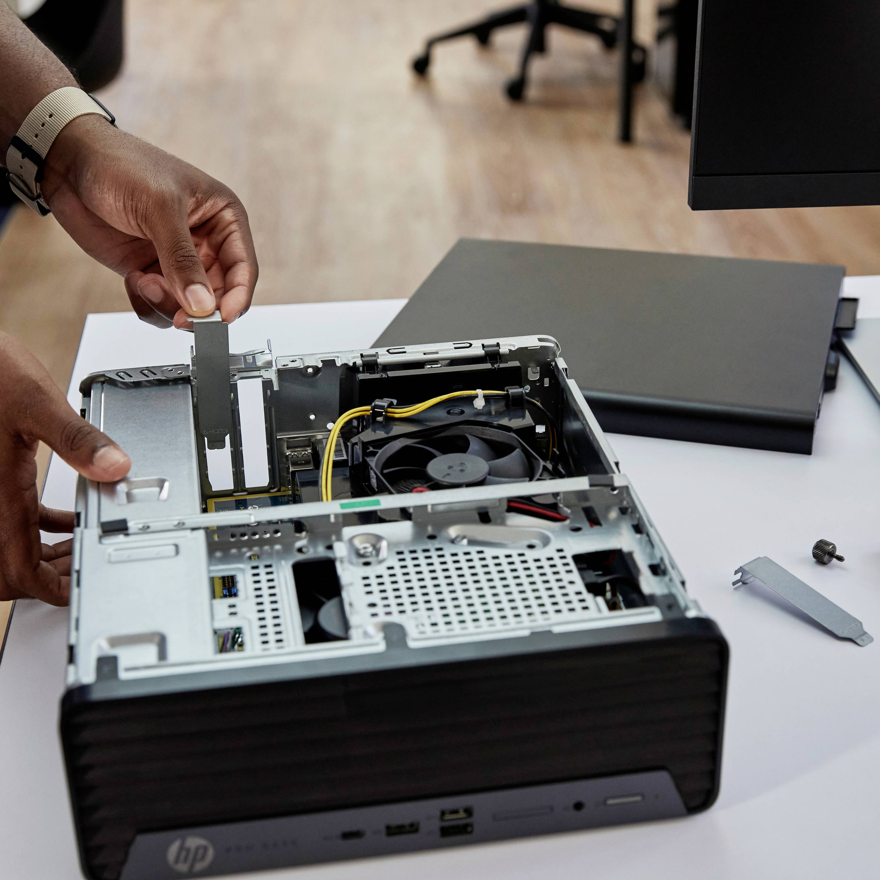 Eine Person installiert eine neue Hardware-Komponente in einen offenen Desktop-Computer, auf einem Schreibtisch mit Monitor im Hintergrund.