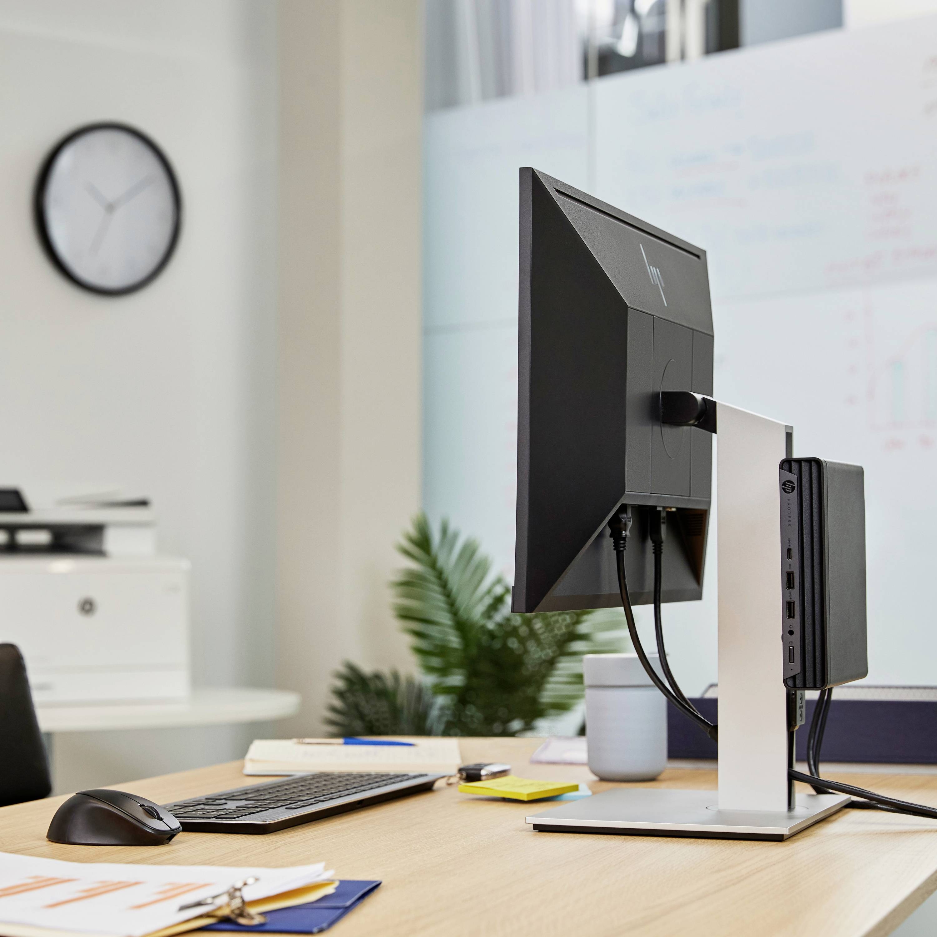 Moderner Büroschreibtisch mit Computer, Monitor, Tastatur, Maus und Notizbüchern; im Hintergrund sind eine Uhr und Pflanzen.