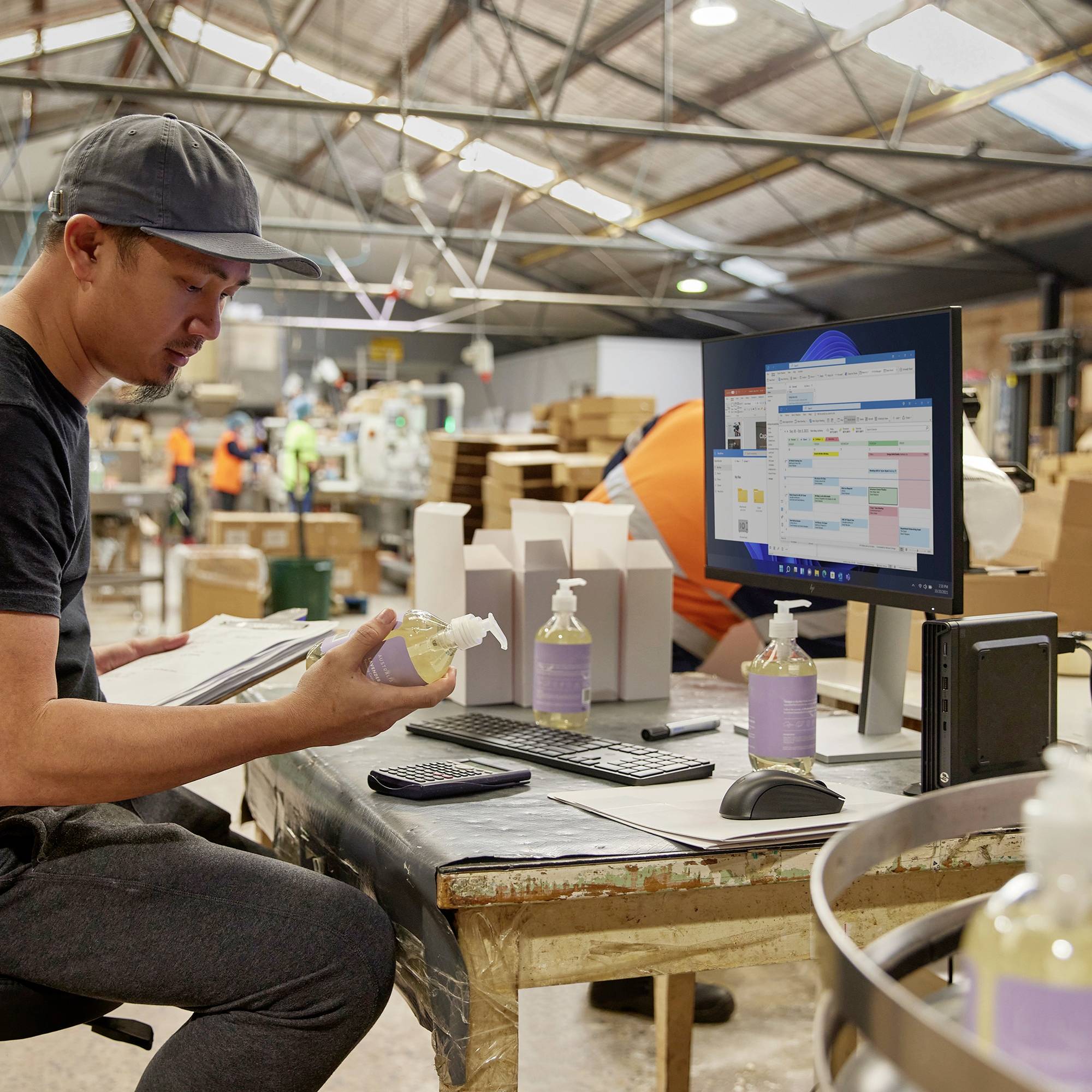 Ein Mann in schwarzem T-Shirt und grauer Kappe arbeitet an einem Holztisch in einer Fabrik. Er überprüft Dokumente und hat ein Handdesinfektionsmittel und eine Rechnungsmaschine zur Hand. Im Hintergrund sind Produktionsmitarbeiter und Kartons zu sehen.