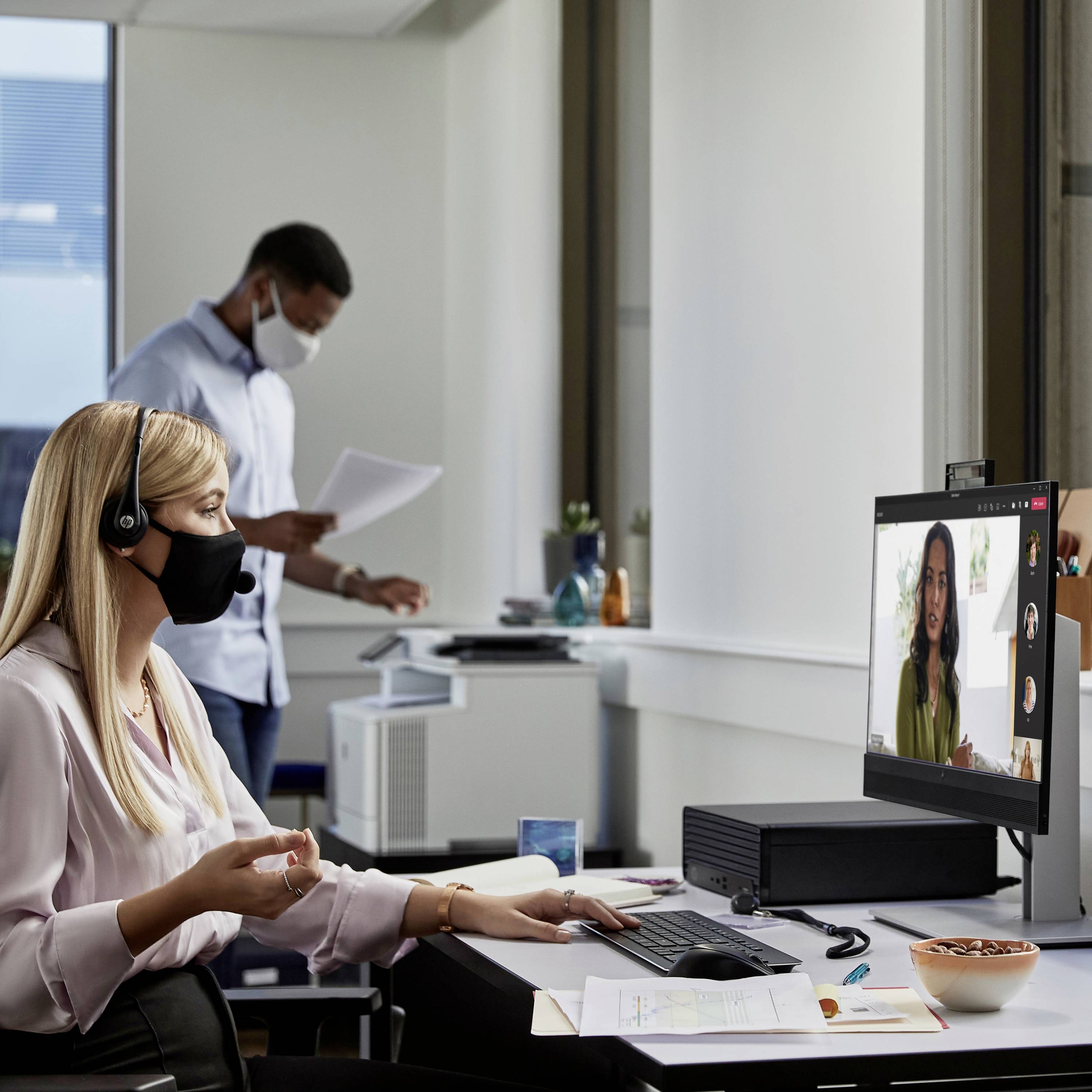 Zwei Personen arbeiten in einem modernen Büro. Eine Frau in einem Videoanruf trägt ein Headset, während ein Mann hinter ihr ein Dokument liest.