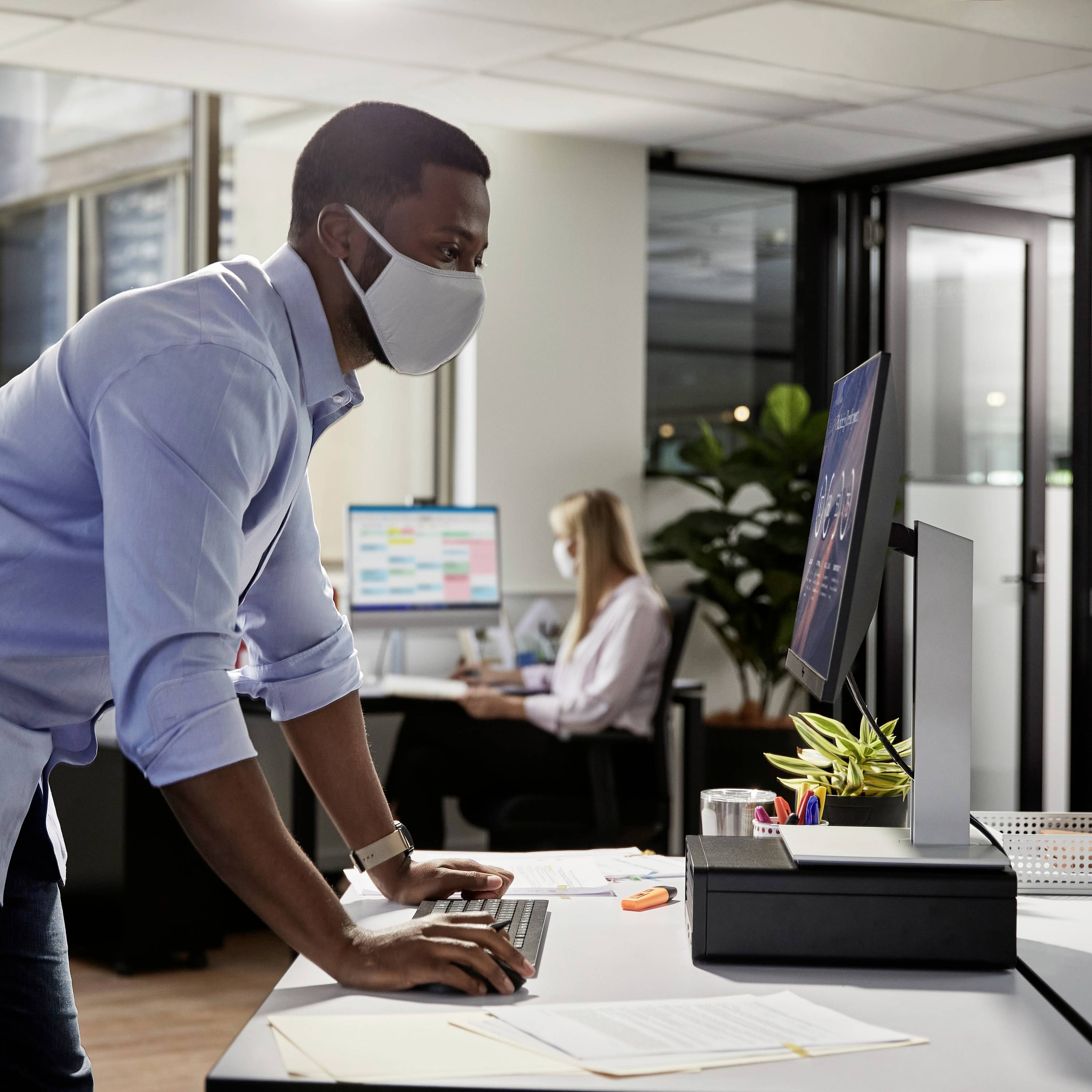 Ein Mann mit Gesichtsmaske arbeitet an einem Computer in einem modernen Büro. Im Hintergrund sitzt eine Frau ebenfalls am Computer.