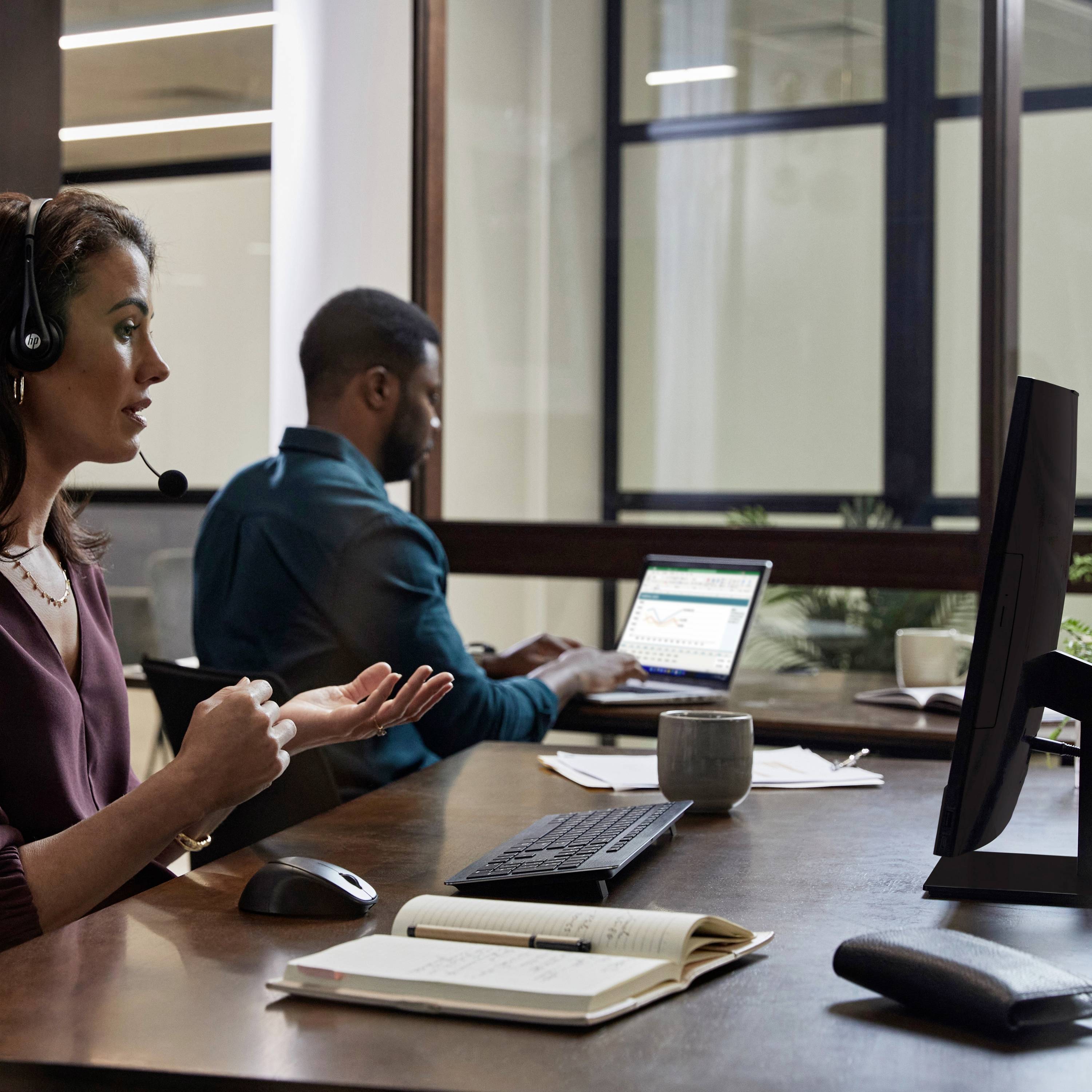 Eine Frau mit Headset sitzt an einem Schreibtisch und gestikuliert, während ein Mann im Hintergrund an einem Laptop arbeitet.