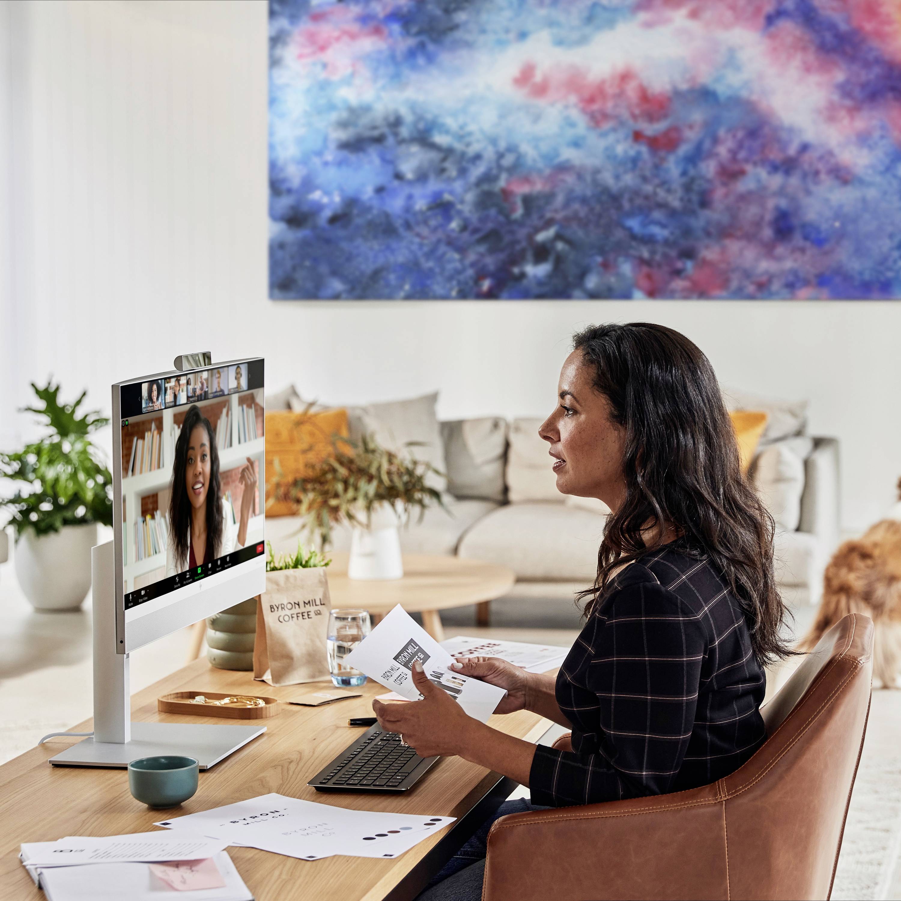 Eine Frau sitzt vor einem Computer in einer Videokonferenz. Ein großer bunter Wandteppich hängt im Hintergrund, ein Hund steht daneben.