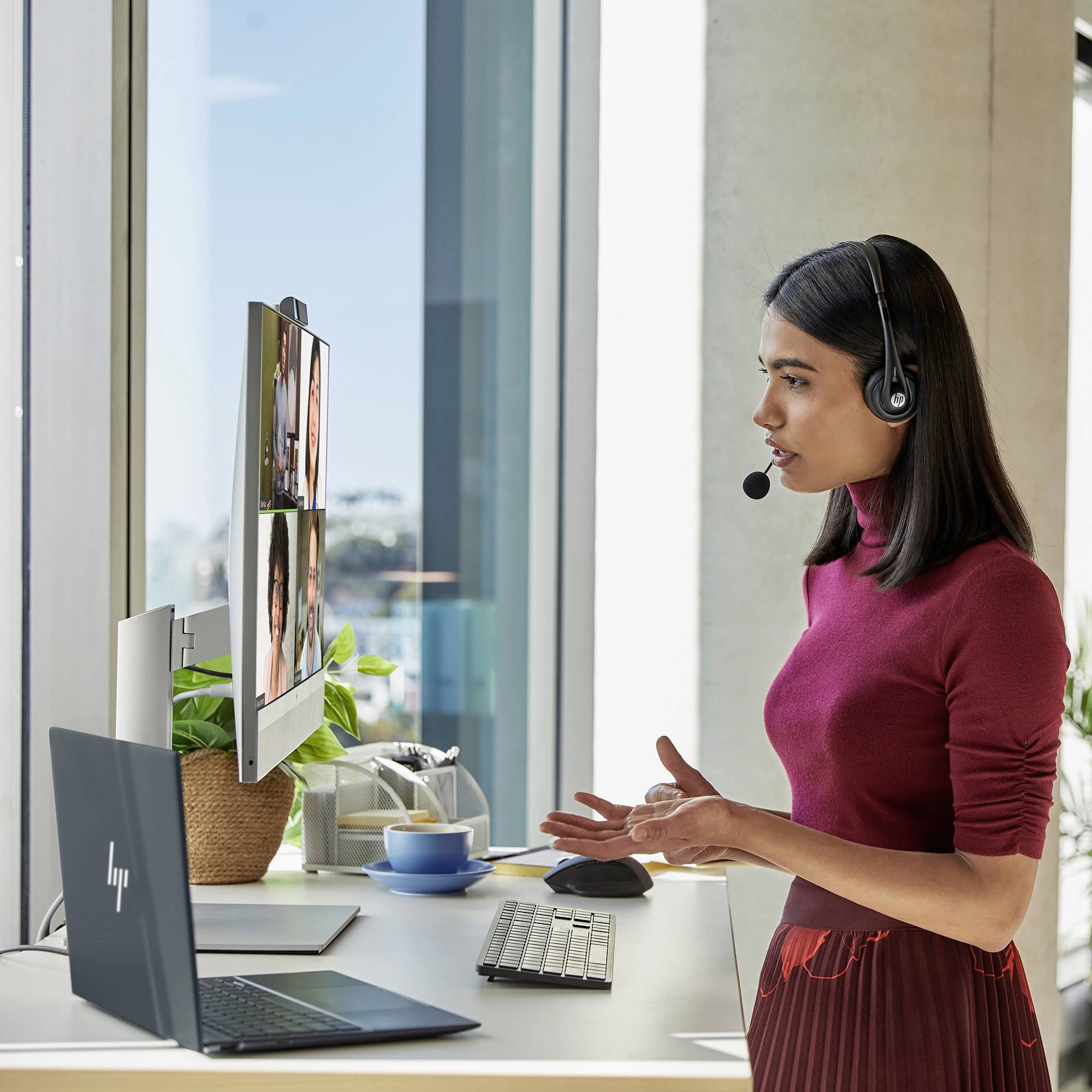 Eine Frau mit Headset steht an einem Schreibtisch und hält eine Videokonferenz. Laptop, Bildschirm, Tastatur und Pflanze sind sichtbar.