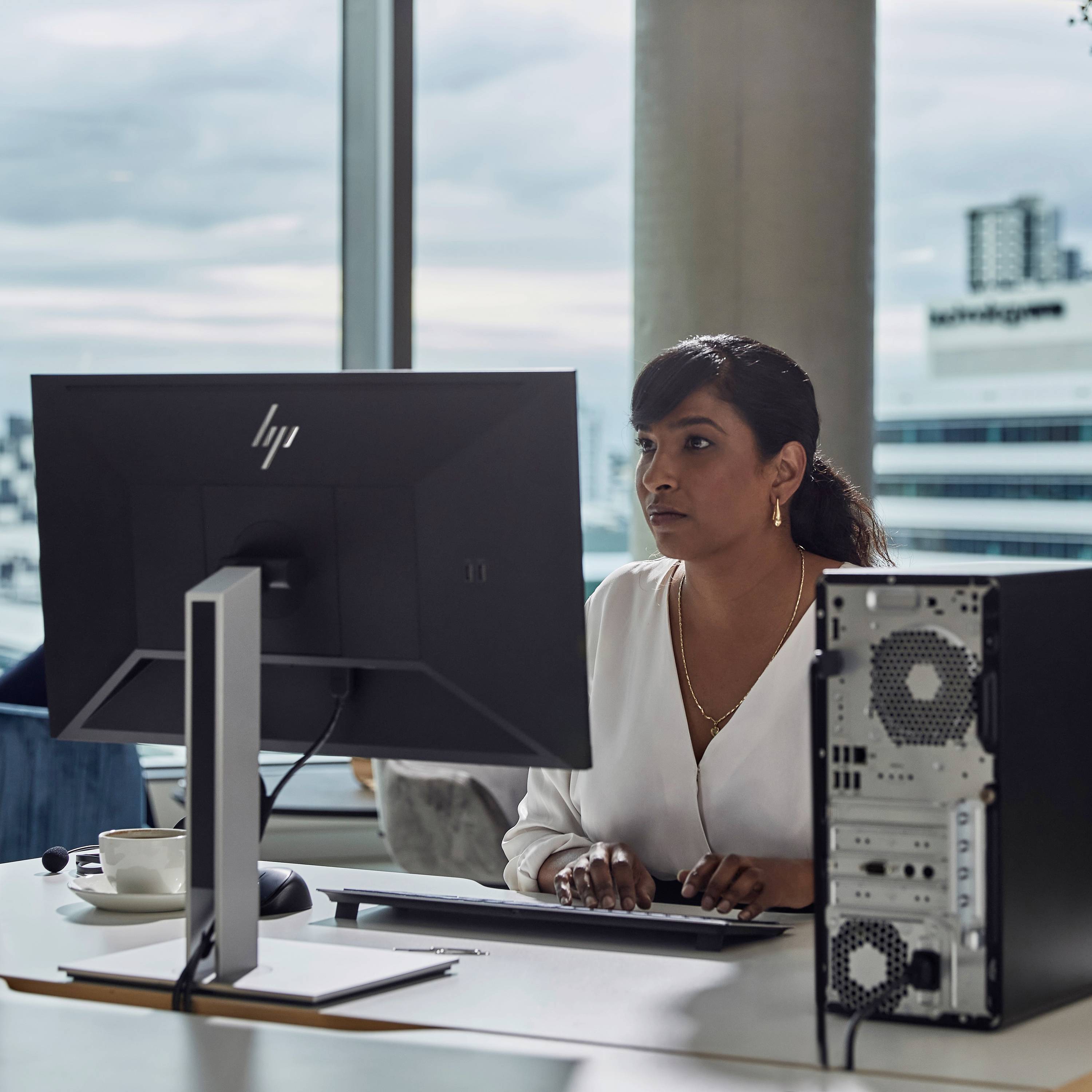 Eine Frau arbeitet an einem Computer in einem modernen Büro mit Blick auf eine Stadtlandschaft. Ein großer Bildschirm und ein PC-Tower sind sichtbar.