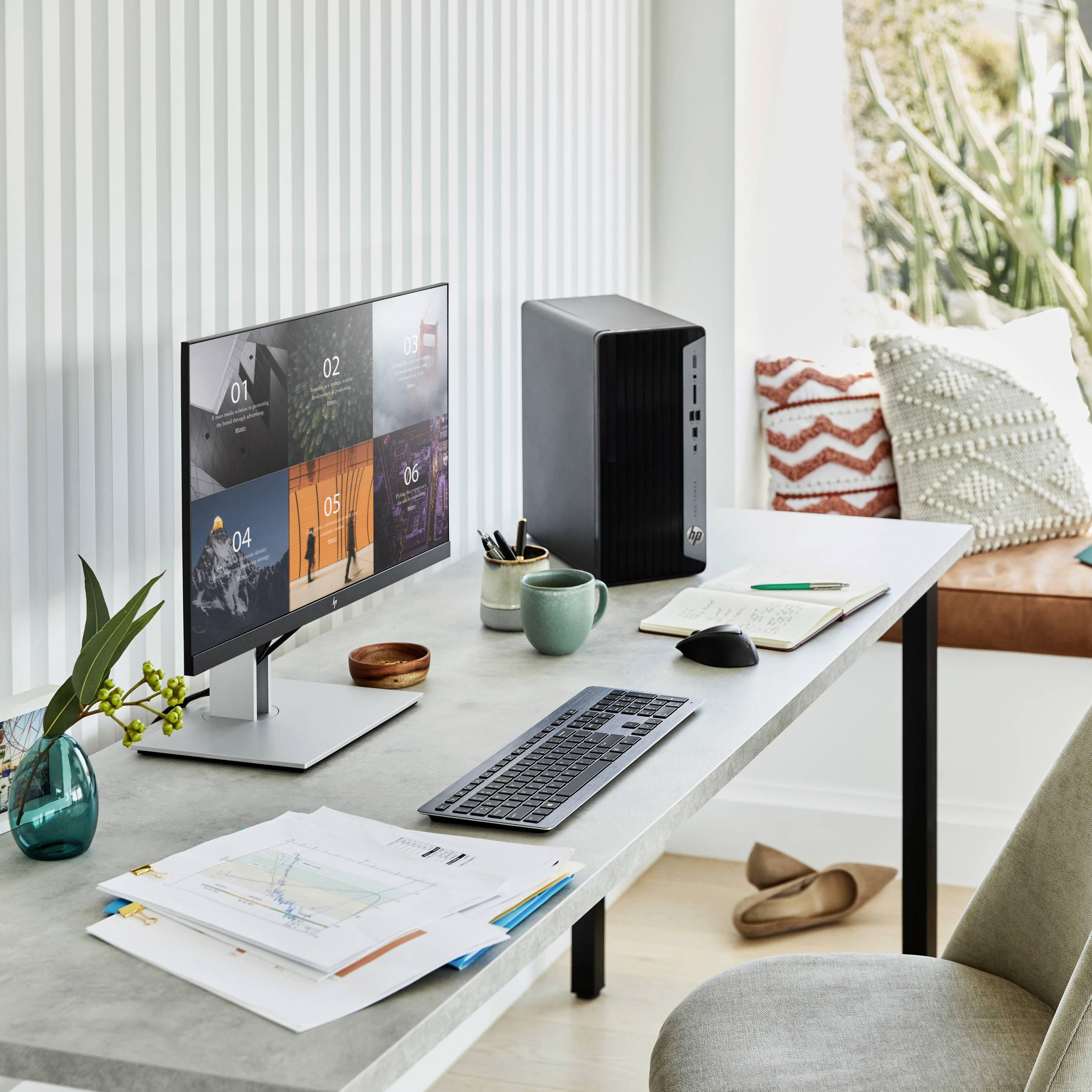 Moderner Arbeitsplatz mit Computer, Tastatur, Maus und Dokumenten. Im Hintergrund Pflanze und Lampe, Fenster mit Tageslicht.