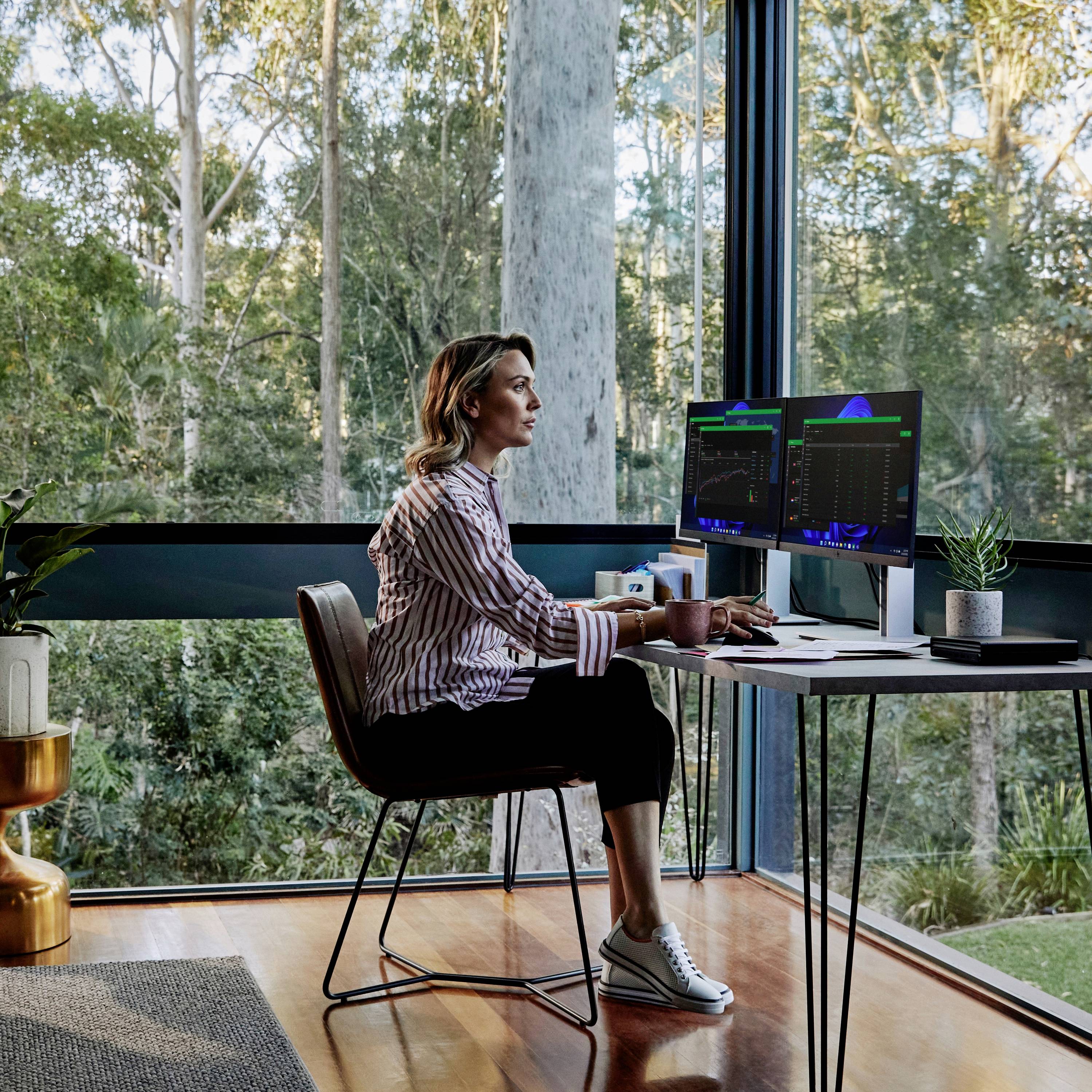 Eine Person arbeitet an einem Computer neben einem großen Fenster mit Blick auf einen Wald.