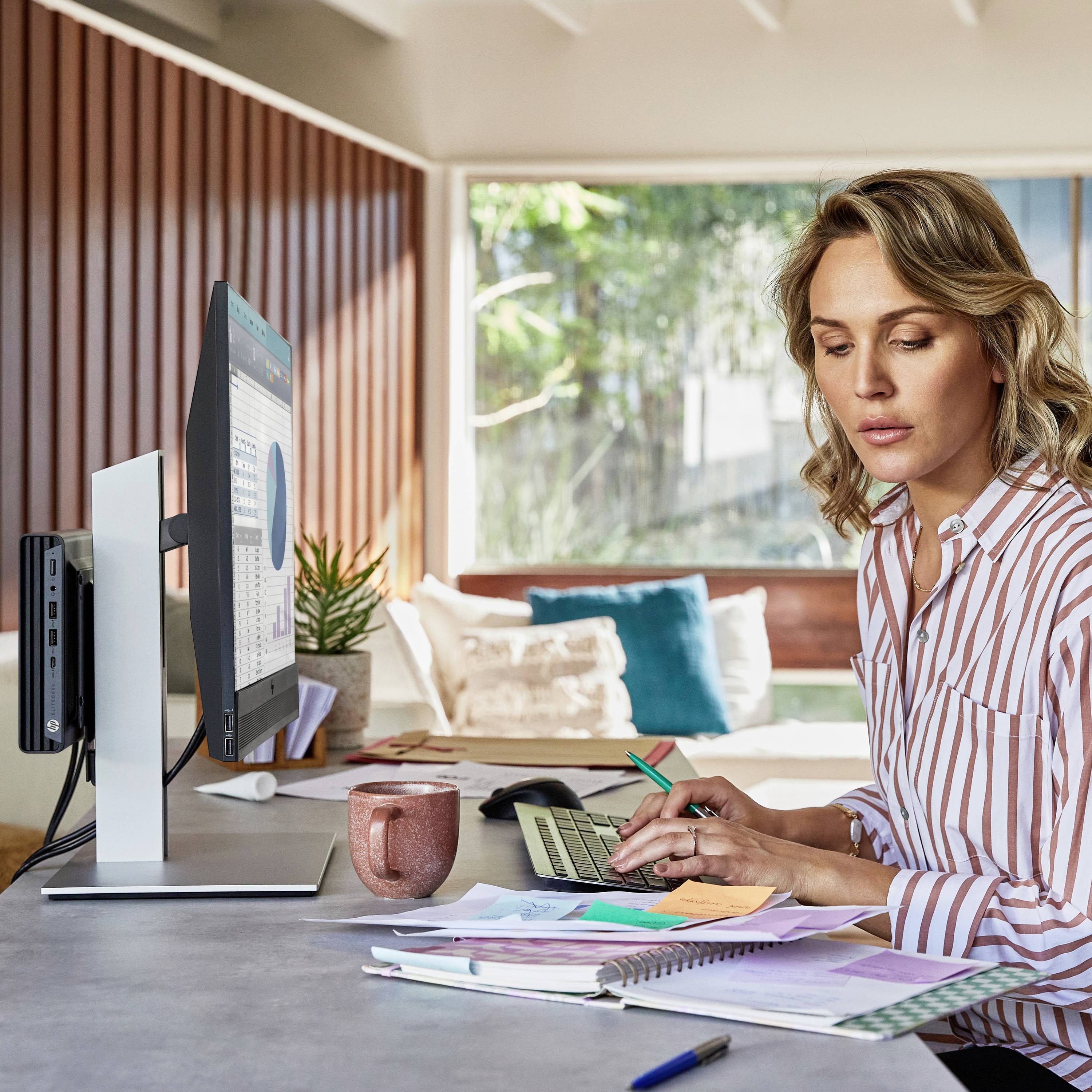 Eine Frau sitzt an einem Schreibtisch und arbeitet an Dokumenten. Ein Computerbildschirm zeigt Tabellen und ein Fenster bietet Tageslicht.