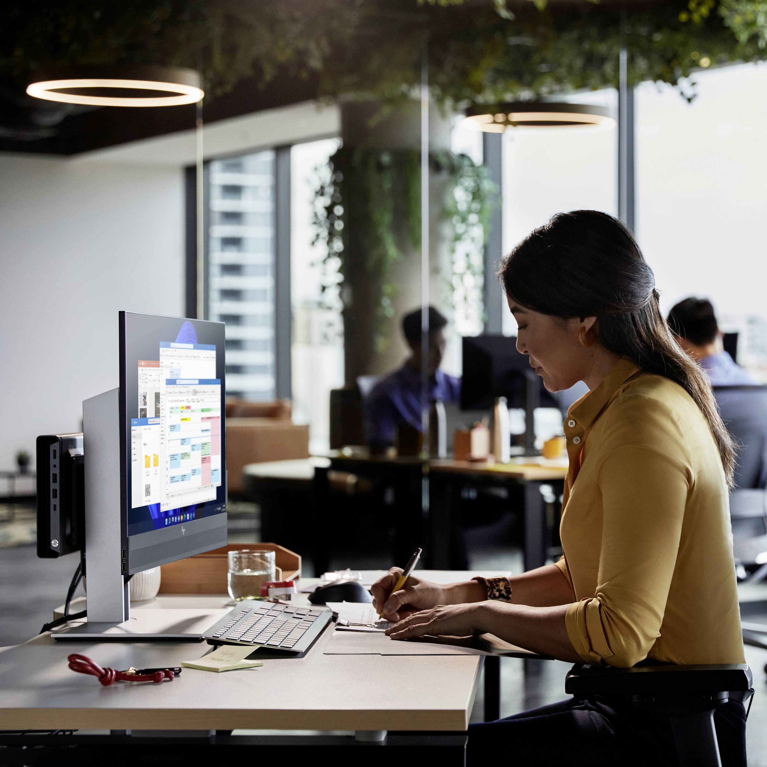 Eine Frau sitzt an einem Schreibtisch in einem modernen Büro, arbeitet am Computer, während Menschen im Hintergrund arbeiten.