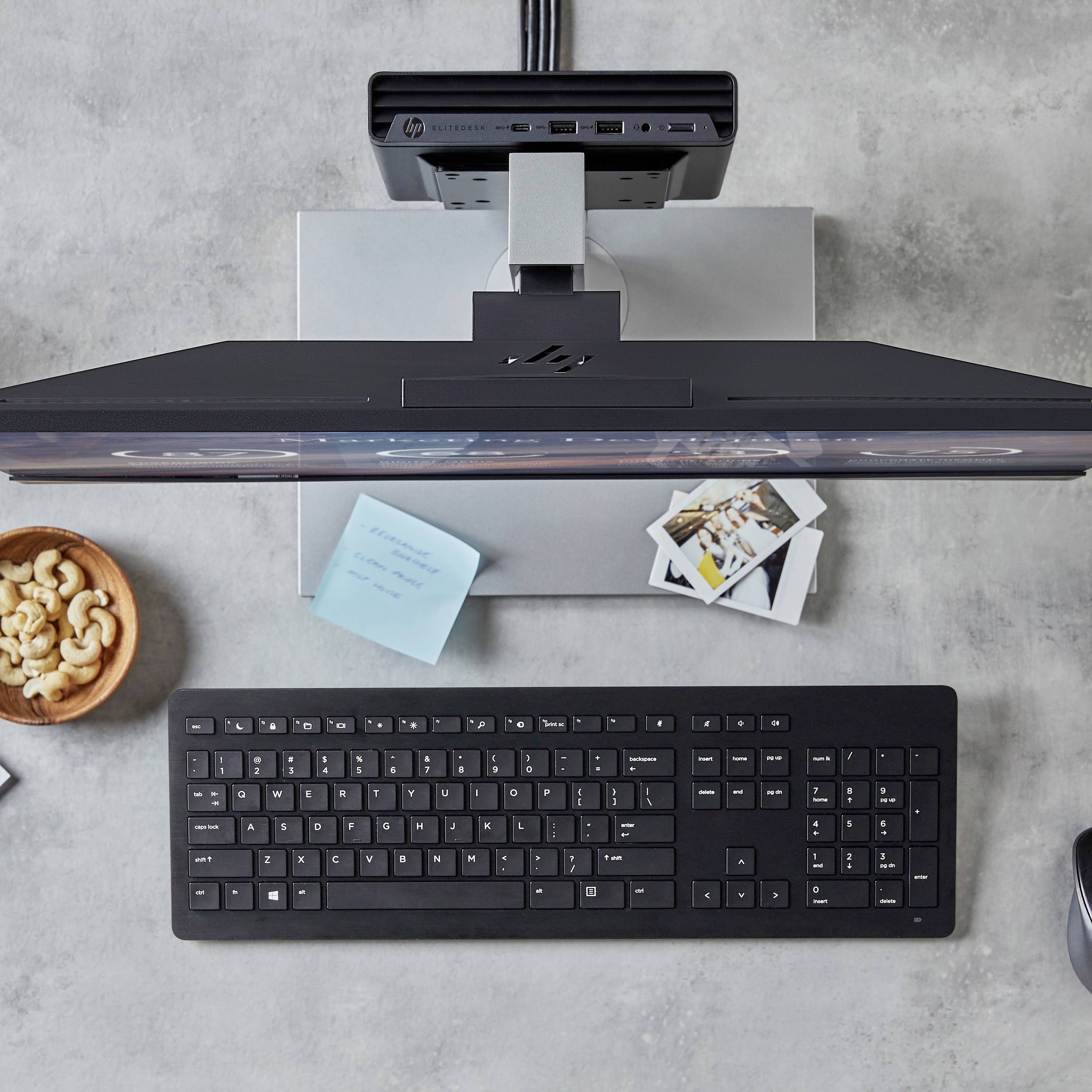 Ein Schreibtisch mit einer Computertastatur, einem Monitor, einem Notizblock, Fotos und einem Teller mit Cashewnüssen.