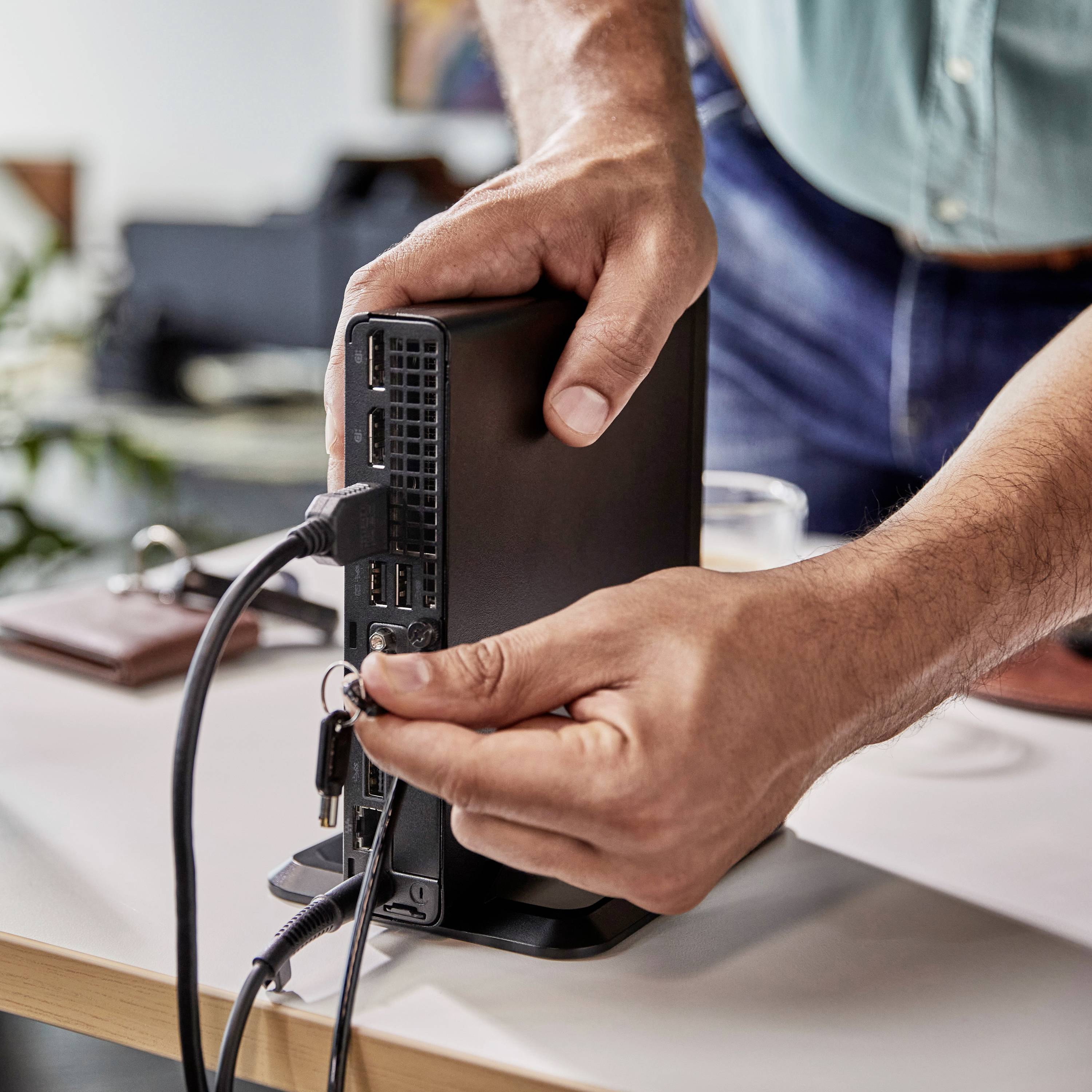 Person verbindet Kabel mit kleiner Computerbox auf Schreibtisch, im Hintergrund Büroeinrichtung erkennbar.
