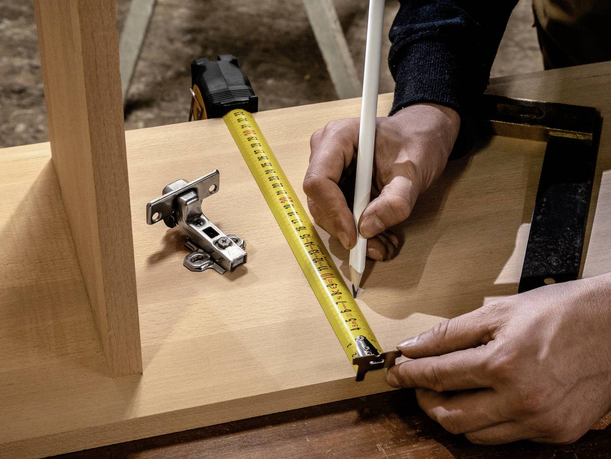 Eine Person misst ein Holzbrett mit einem Maßband und markiert es mit einem Stift. Ein Scharnier liegt daneben.