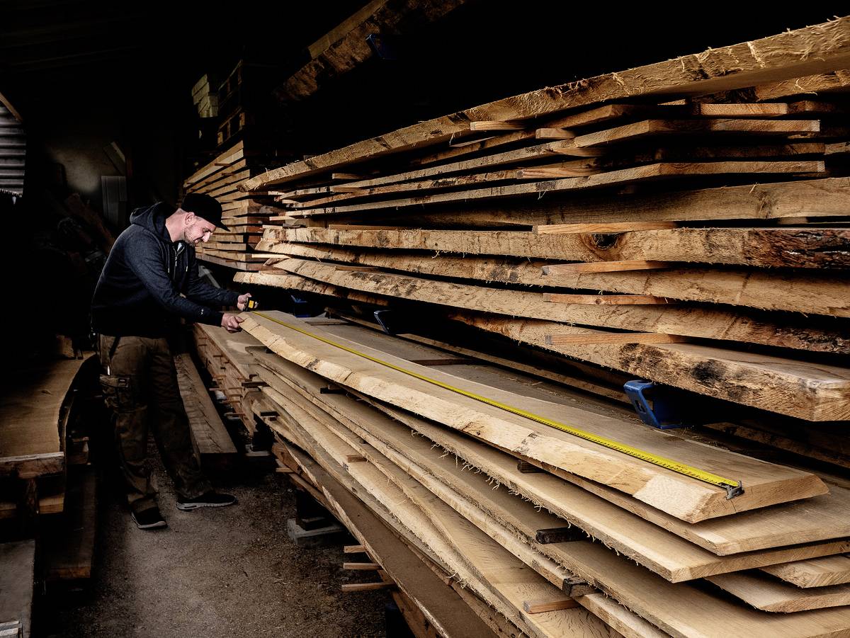 Ein Mann misst in einer Lagerhalle große Holzplanken mit einem Maßband. Im Hintergrund sind gestapelte Holzbretter.