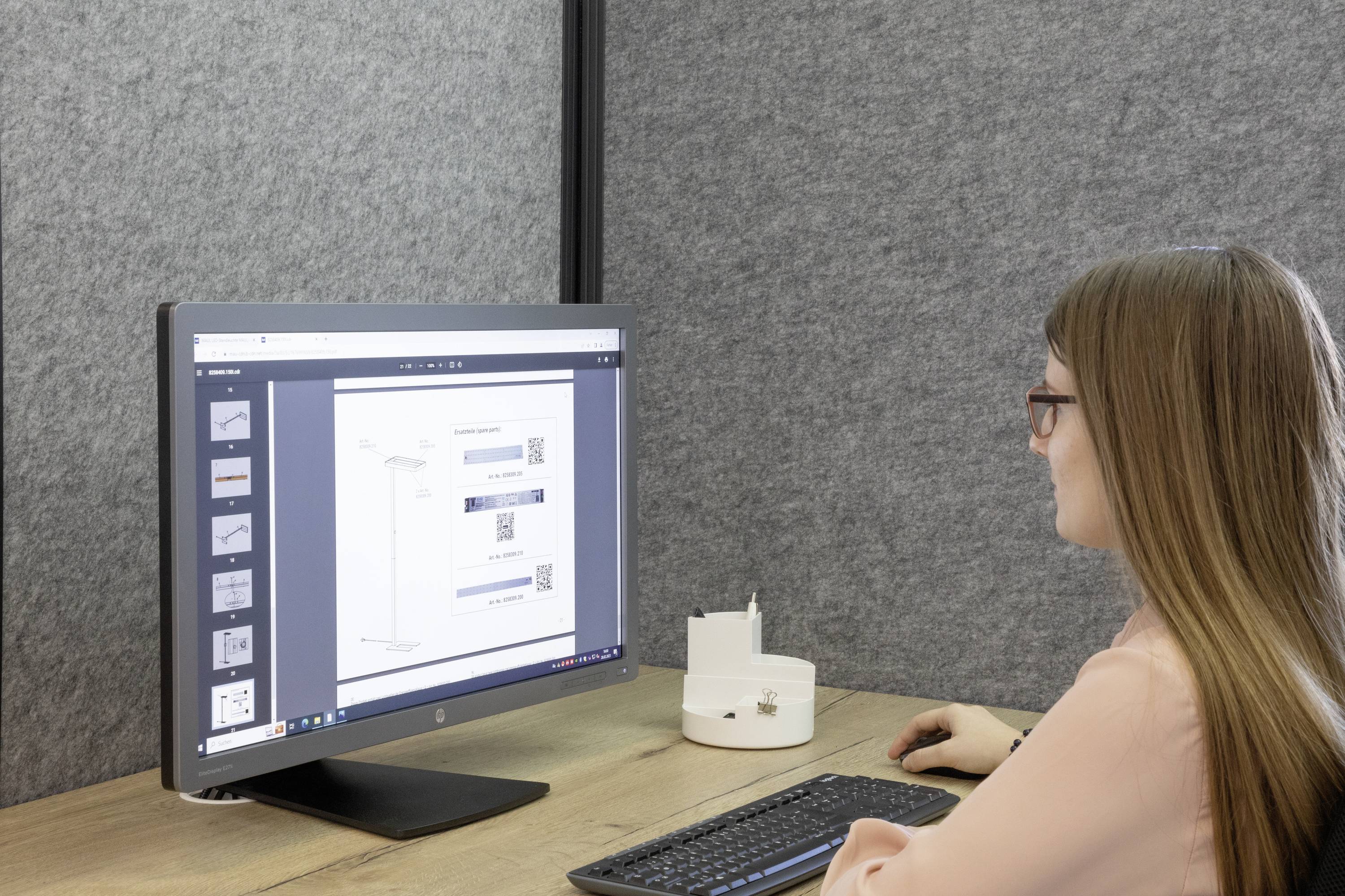 Eine Frau sitzt am Schreibtisch und betrachtet ein Dokument auf einem Computermonitor. Sie trägt eine Brille und hat langes Haar.