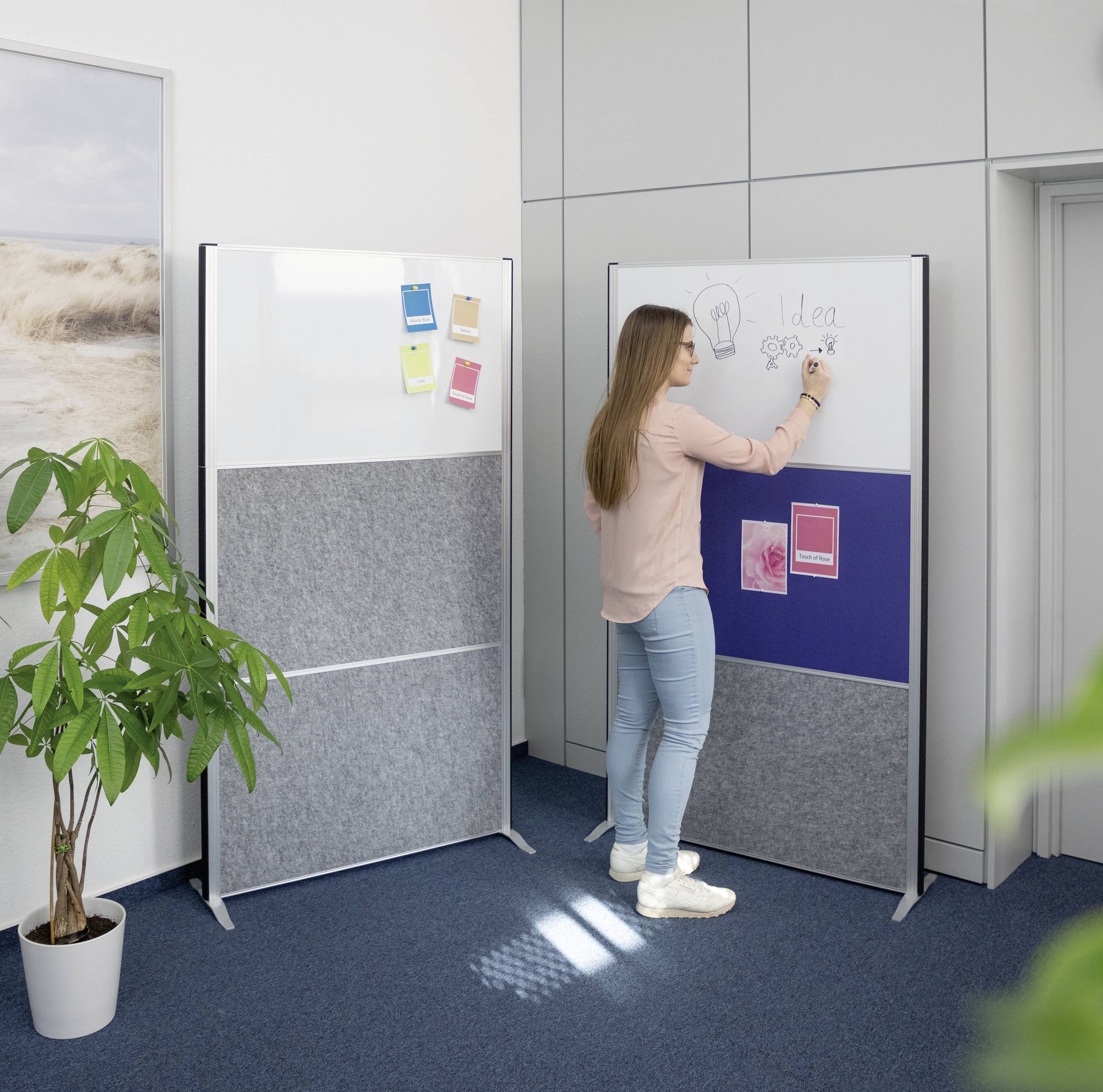 Eine Frau schreibt auf eine große Pinnwand in einem Büro. Links steht eine Pflanze, im Hintergrund ist eine weitere Pinnwand zu sehen.