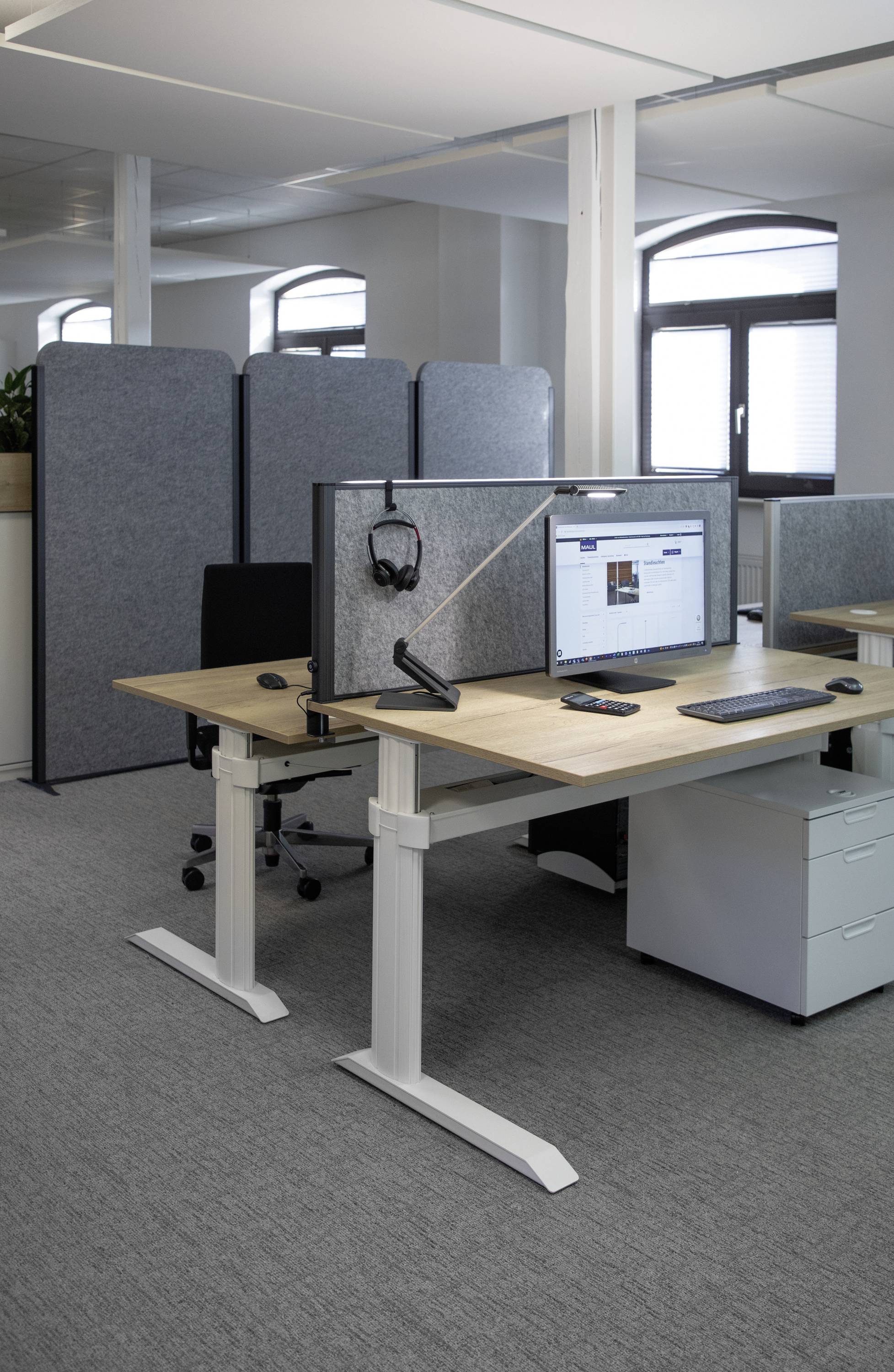 Ein modernes Büro mit höhenverstellbaren Schreibtischen, Bildschirm und Laptops. Sichtschutzwände trennen die Arbeitsplätze.