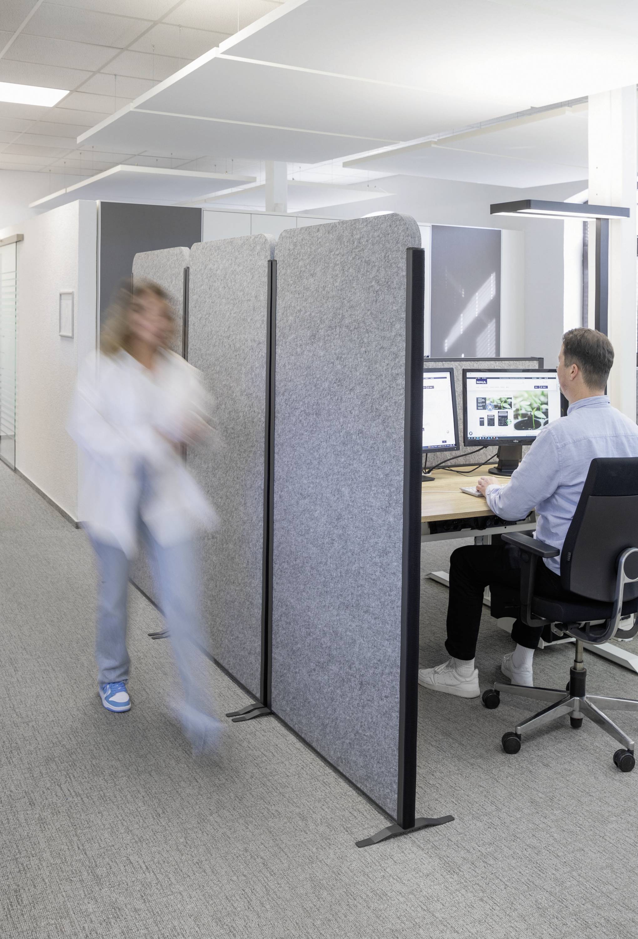 Ein Mann sitzt am Schreibtisch und arbeitet am Computer. Eine Frau geht schnell vorbei. Büro mit modernem Design und Trennwand.