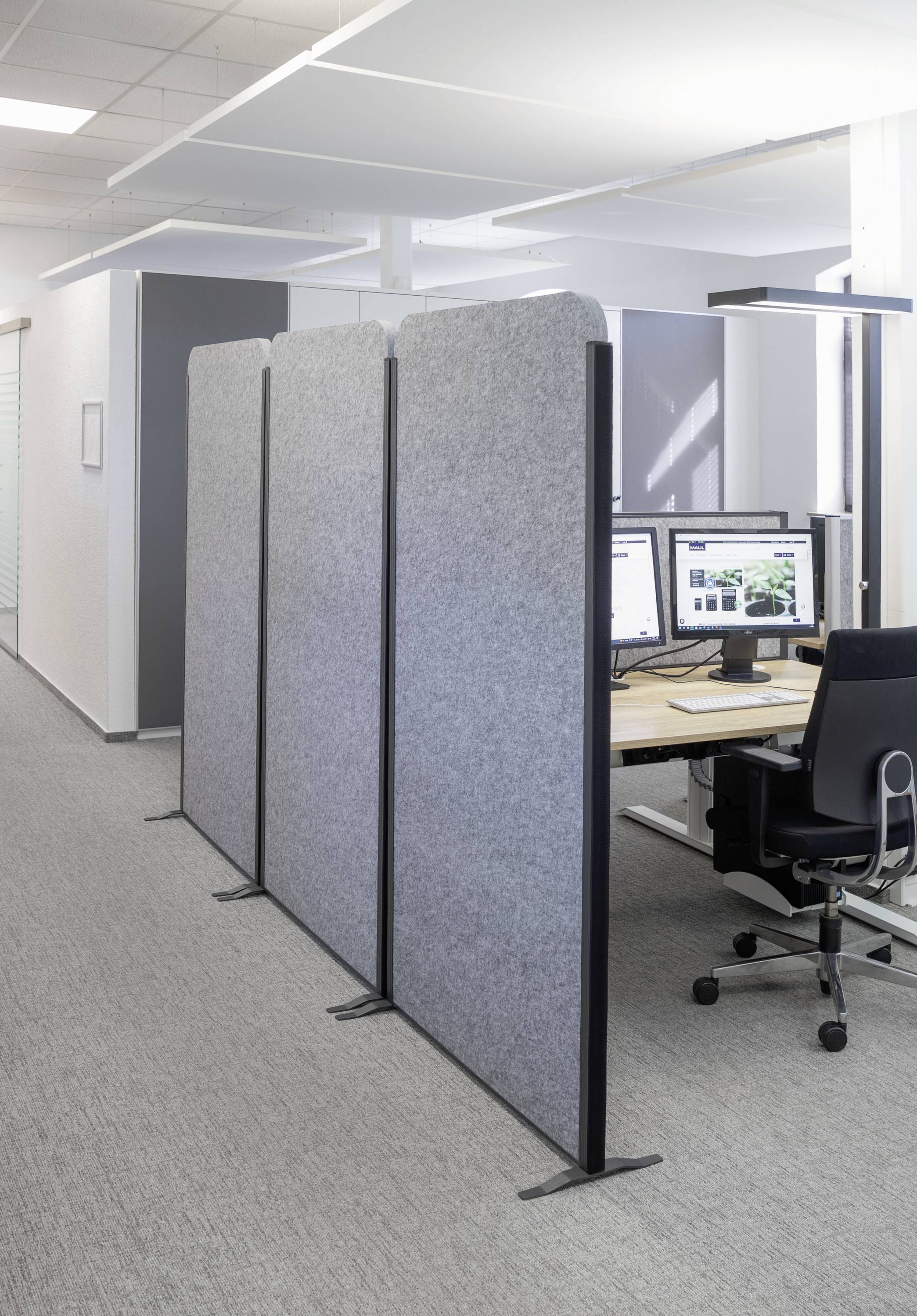 Offenes Büro mit grauen Akustiktrennwänden, die Arbeitsplätze für Privatsphäre teilen. Im Hintergrund sind Schreibtische mit Computermonitoren.