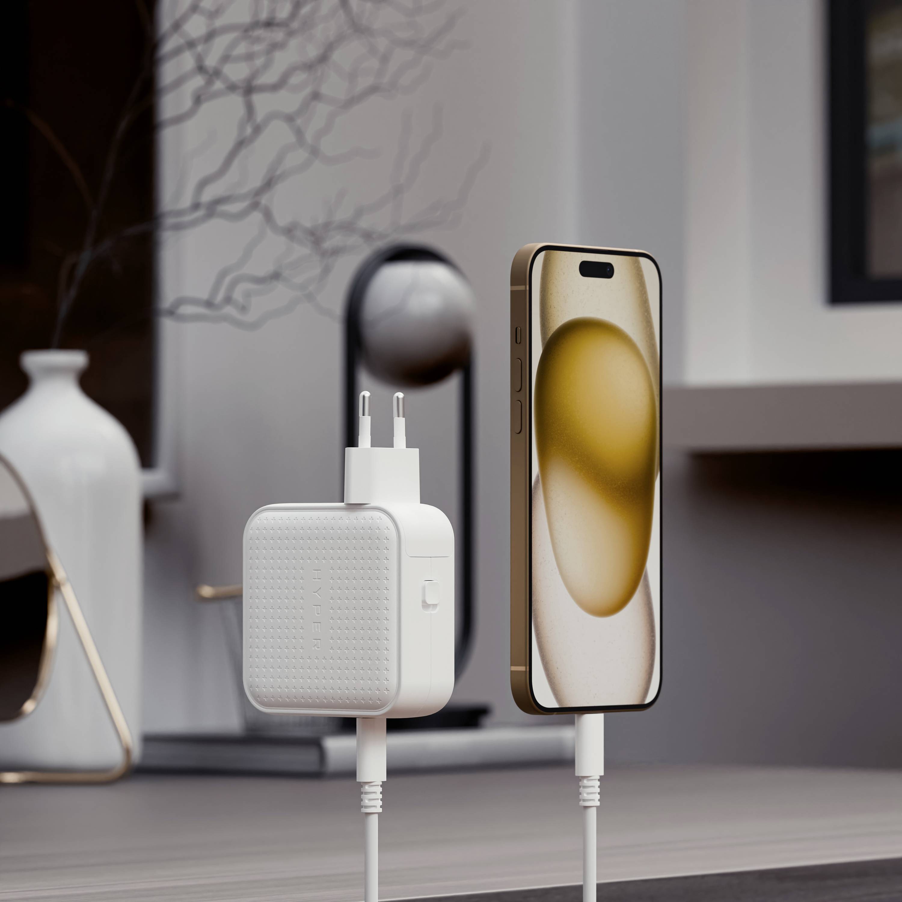 Ein Smartphone steht aufrecht neben einem geladenen Netzteil auf einem Tisch, im Hintergrund ist eine dekorative Vase sichtbar.