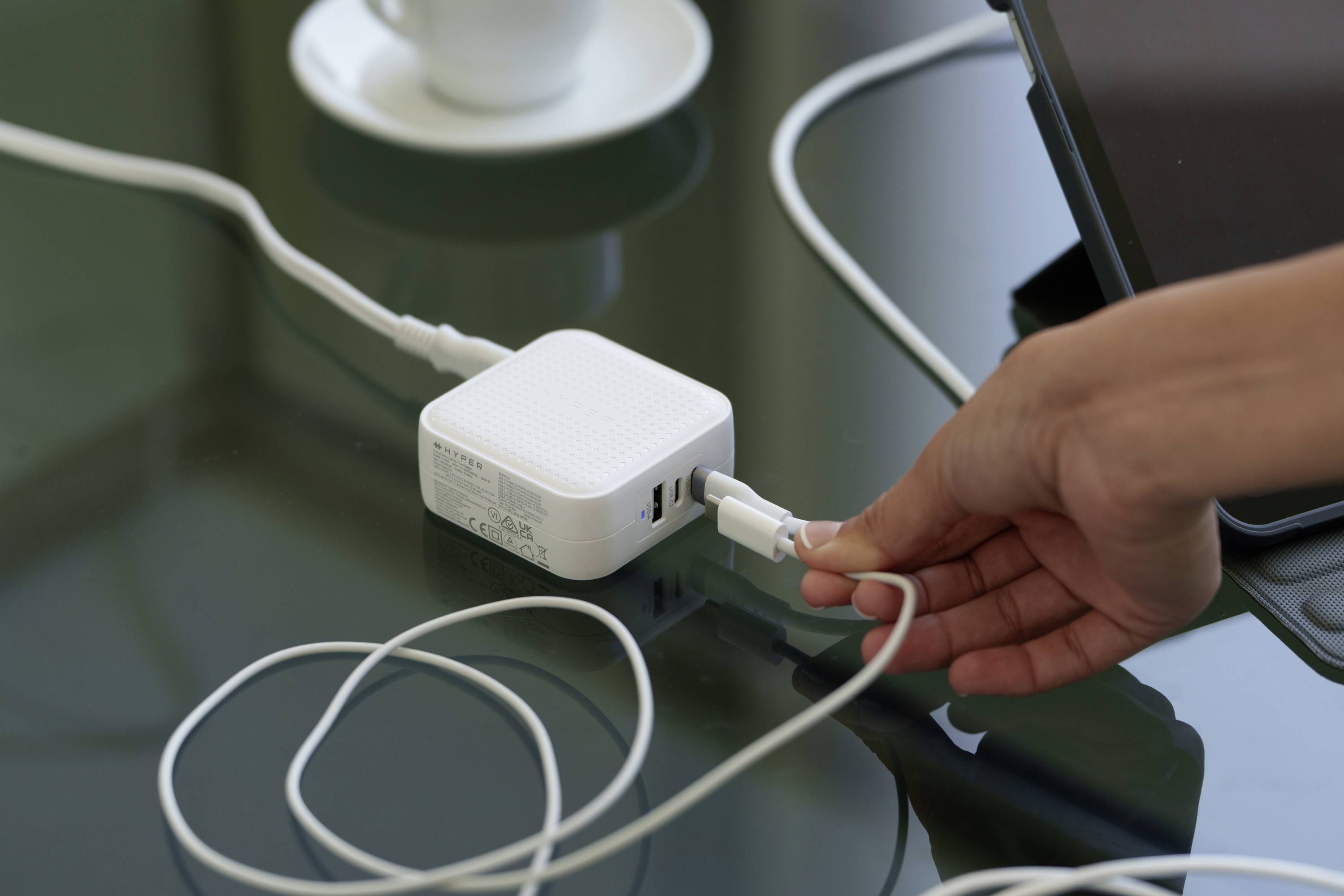 Eine Hand steckt ein Kabel in ein weißes Ladegerät auf einem Tisch. Im Hintergrund ist eine Tasse und ein Tablet sichtbar.