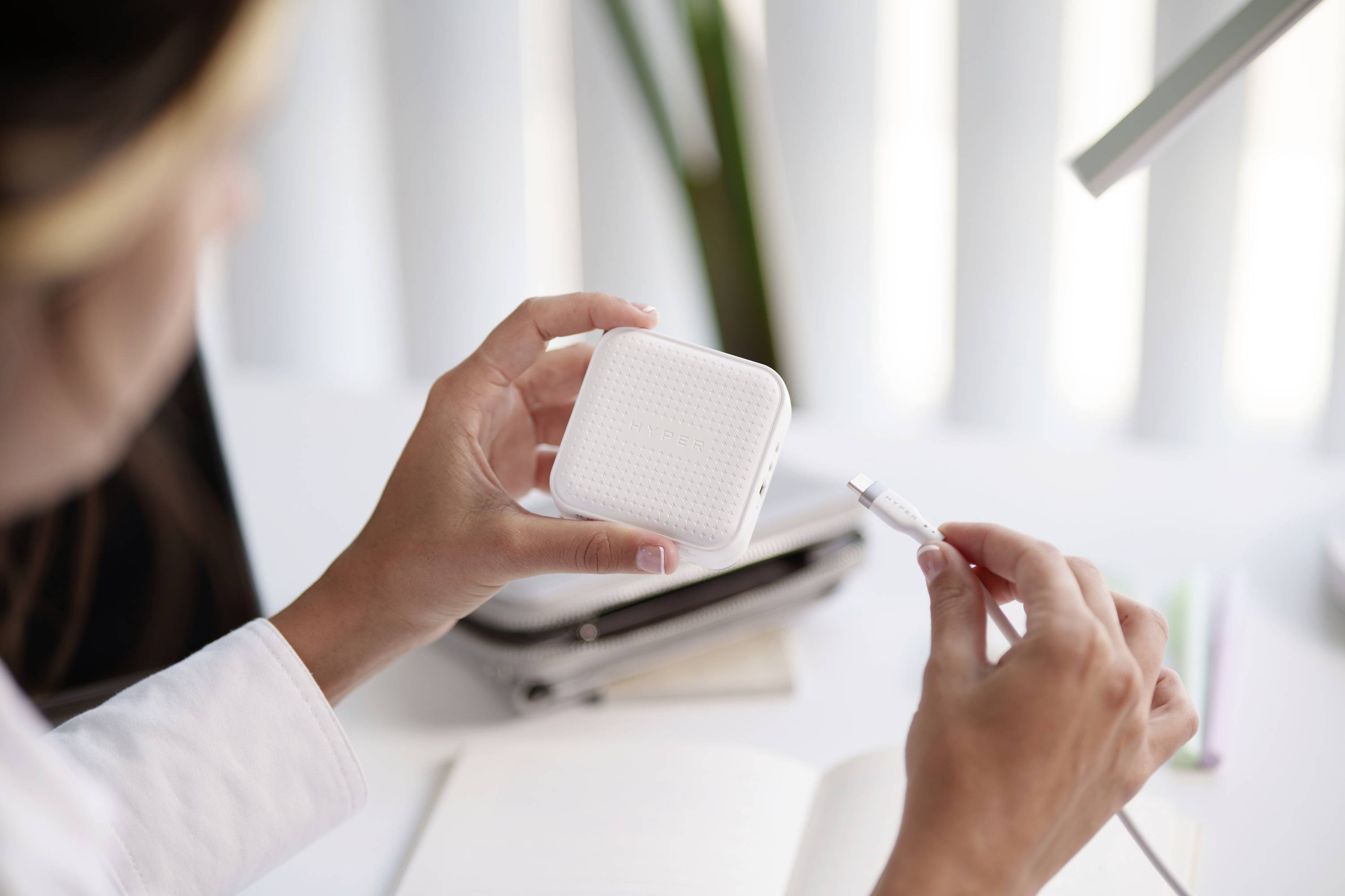 Eine Person hält ein quadratisches elektronisches Gerät und ein Ladekabel in der Hand, sitzend an einem Schreibtisch mit Notizbuch.