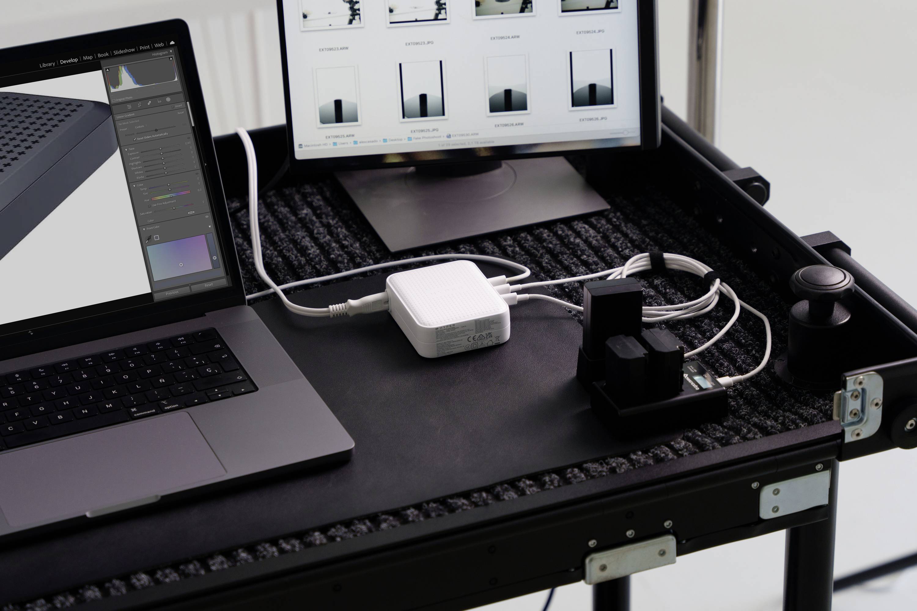 Arbeitsplatz mit Laptop und Monitor auf einem Tisch mit Ladestation und Kabeln. Monitor zeigt Grafiken und Fotobearbeitungssoftware.