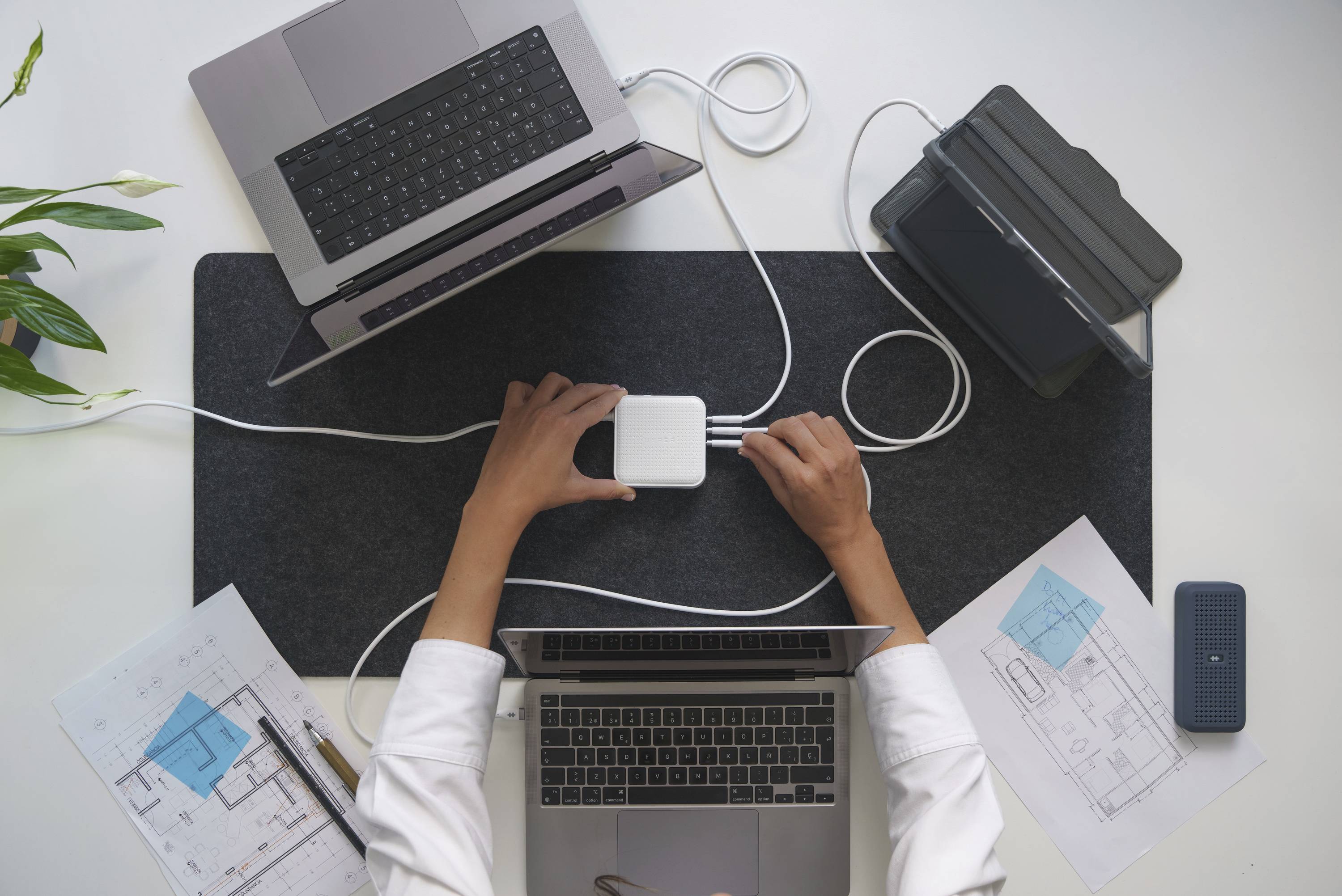 Zwei Laptops, ein Tablet und technische Zeichnungen auf einem Schreibtisch; eine Person verbindet Geräte mit einem Ladegerät.