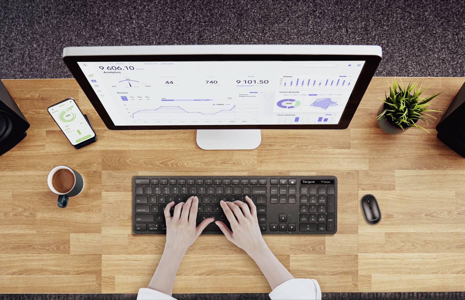 Überblick von oben: Person tippt auf Tastatur vor Monitor mit Diagrammen und Statistiken; Smartphone, Tasse Kaffee und Pflanze daneben.