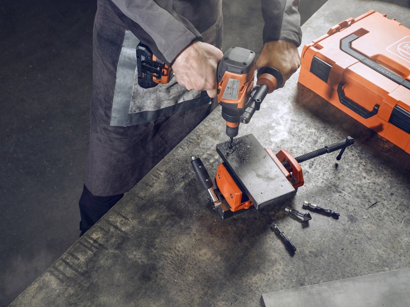 Eine Person bohrt mit einer elektrischen Bohrmaschine in eine Metallplatte, die auf einem Schraubstock befestigt ist. Nebenbei liegt ein orangefarbener Werkzeugkoffer.