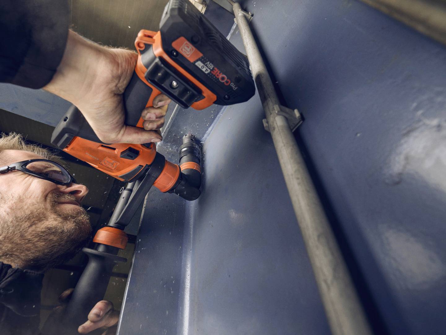 Ein Mann trägt eine Schutzbrille und benutzt eine orange-schwarze Bohrmaschine, um in eine Metallfläche zu bohren.