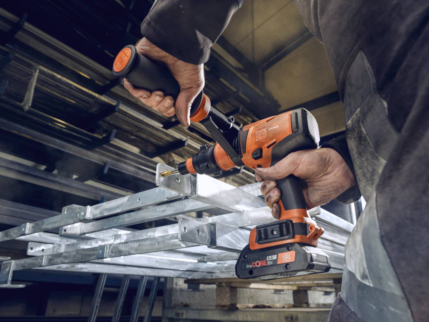 Eine Person verwendet eine orange-schwarze Bohrmaschine, um ein Metallregal in einer industriellen Umgebung zu montieren oder zu bearbeiten.