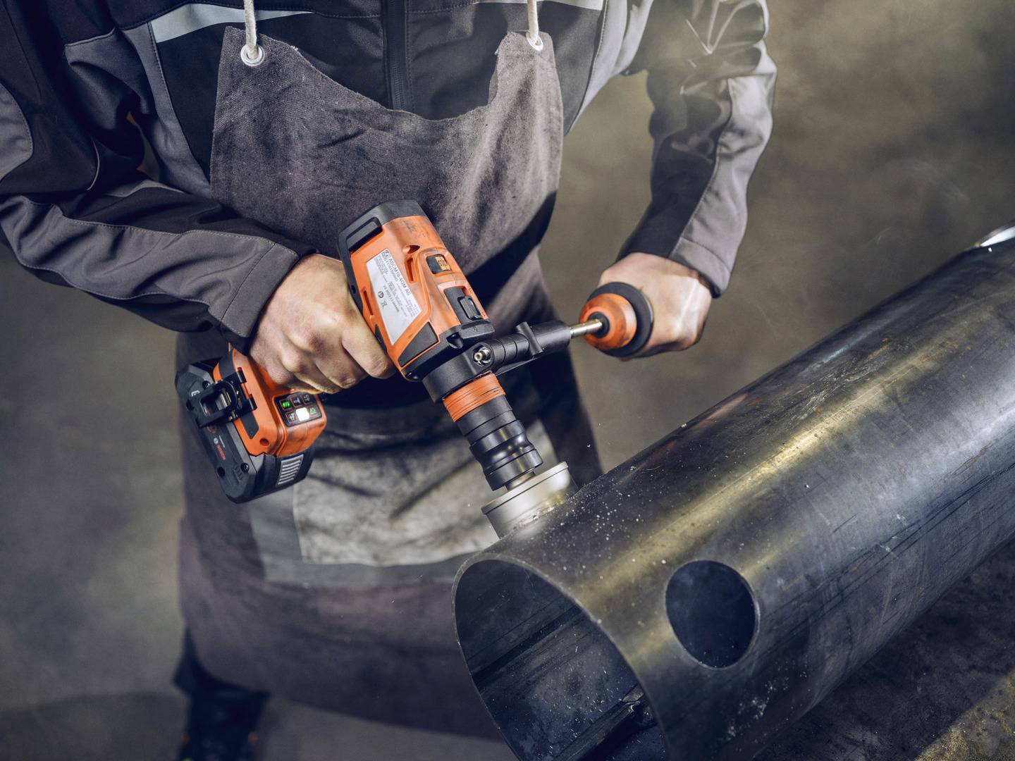 Eine Person in Arbeitskleidung nutzt eine orangefarbene Bohrmaschine, um ein großes Metallrohr zu bearbeiten.