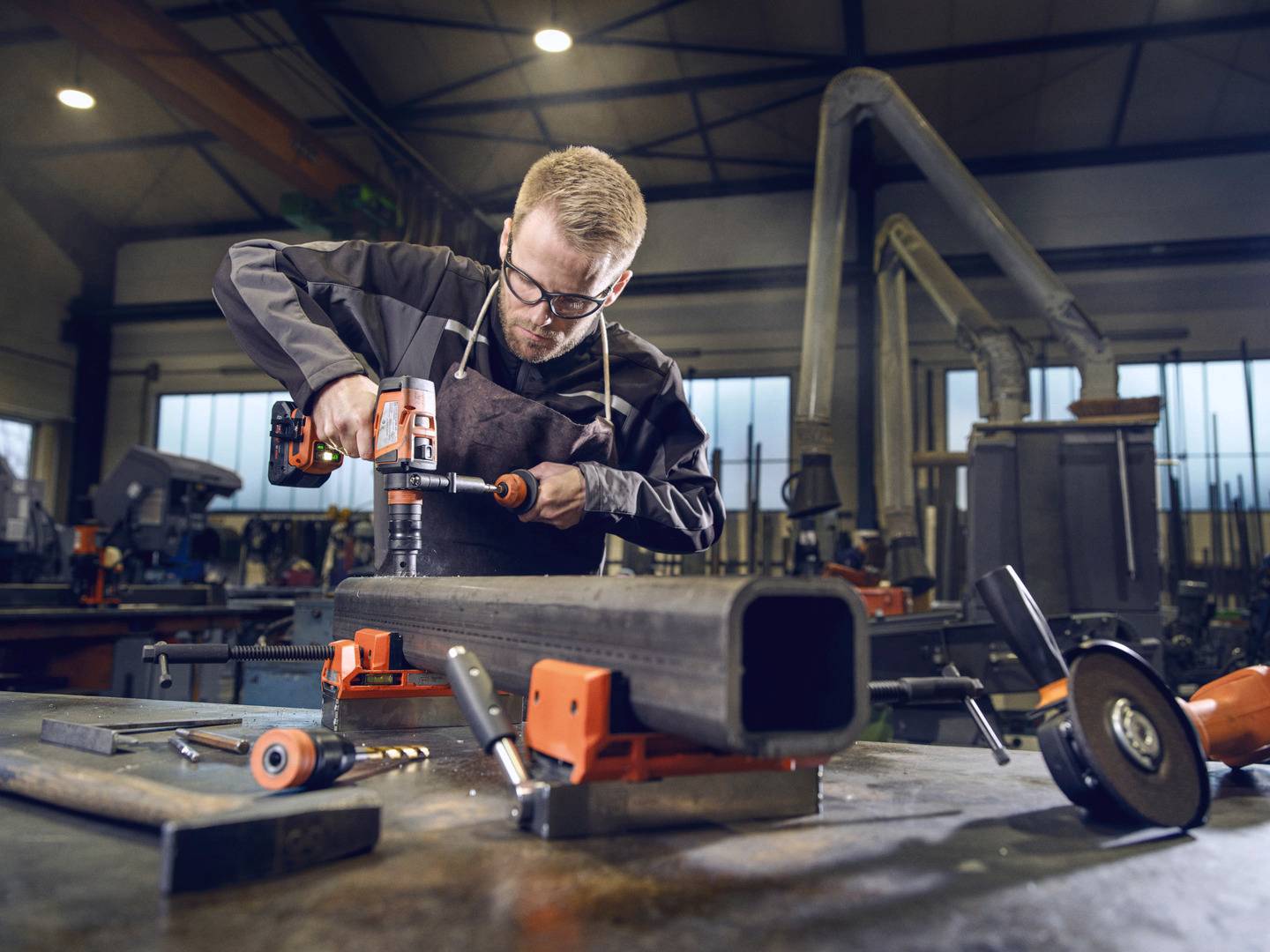 Ein Mann in Schutzkleidung bearbeitet in einer Werkstatt ein großes Metallrohr mit einer Bohrmaschine. Im Hintergrund sind Werkzeuge zu sehen.