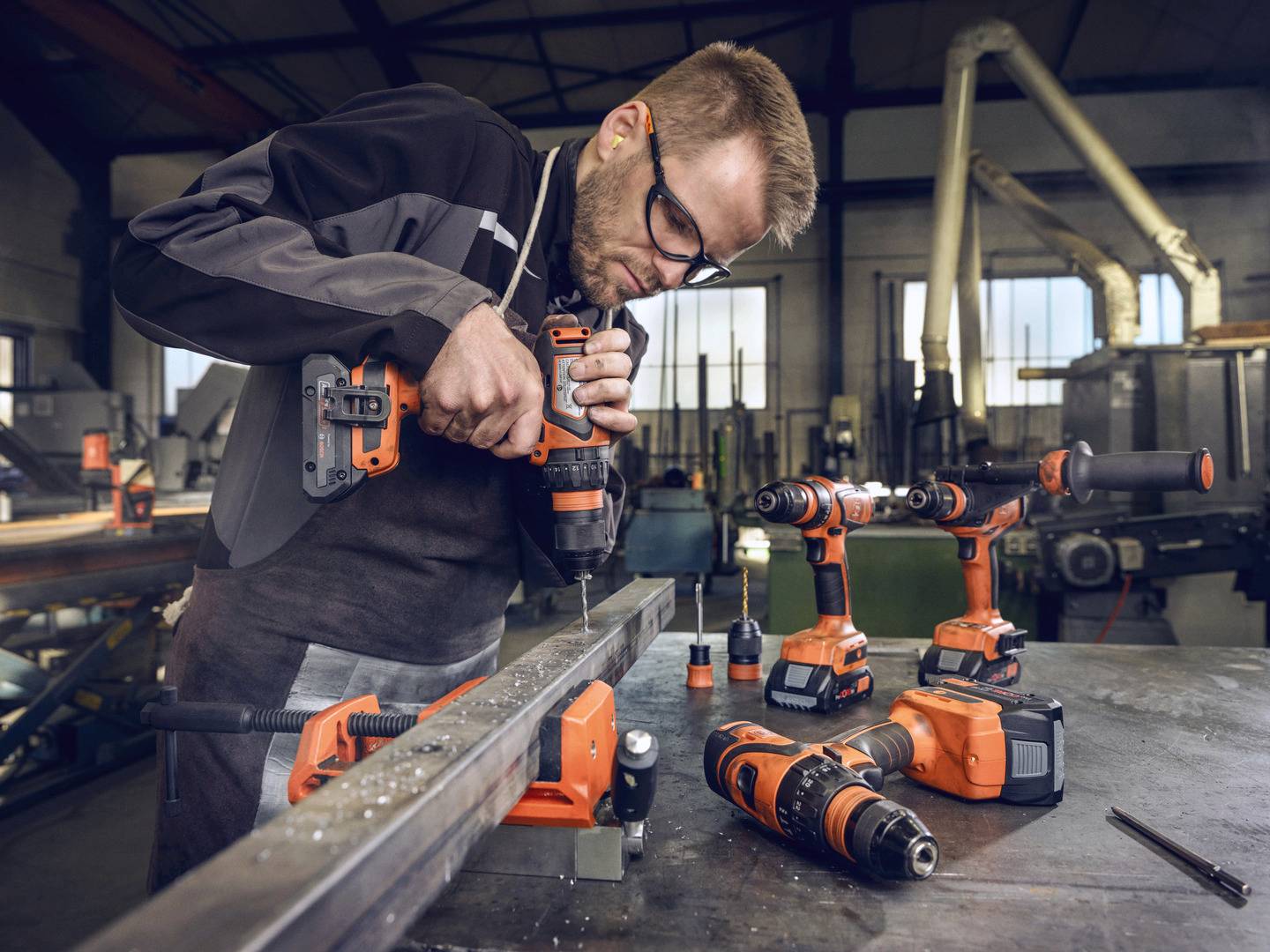 Ein Mann in Arbeitskleidung nutzt eine Bohrmaschine, um in einer metallverarbeitenden Werkstatt ein Metallstück zu bearbeiten.