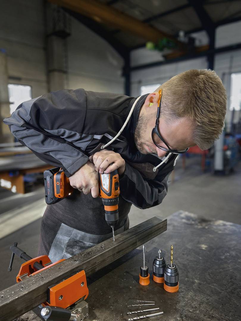 Mann in Werkstatt bohrt mit einer elektrischen Bohrmaschine in ein Metallstück. Schutzbrille und Arbeitskleidung sind sichtbar.