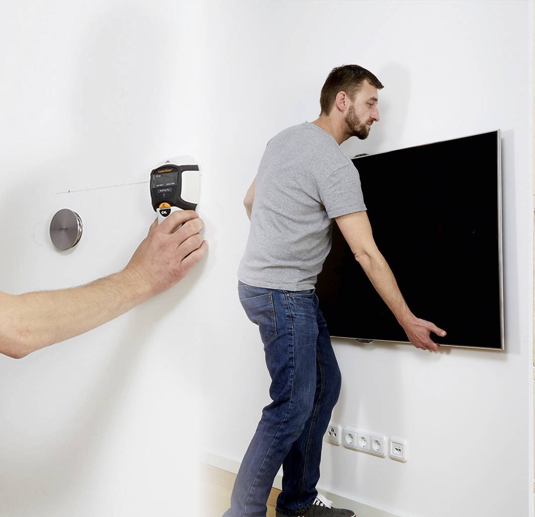 Ein Mann hängt einen Flachbildfernseher an die Wand. Im Hintergrund ist ein Maßband an der Wand befestigt.