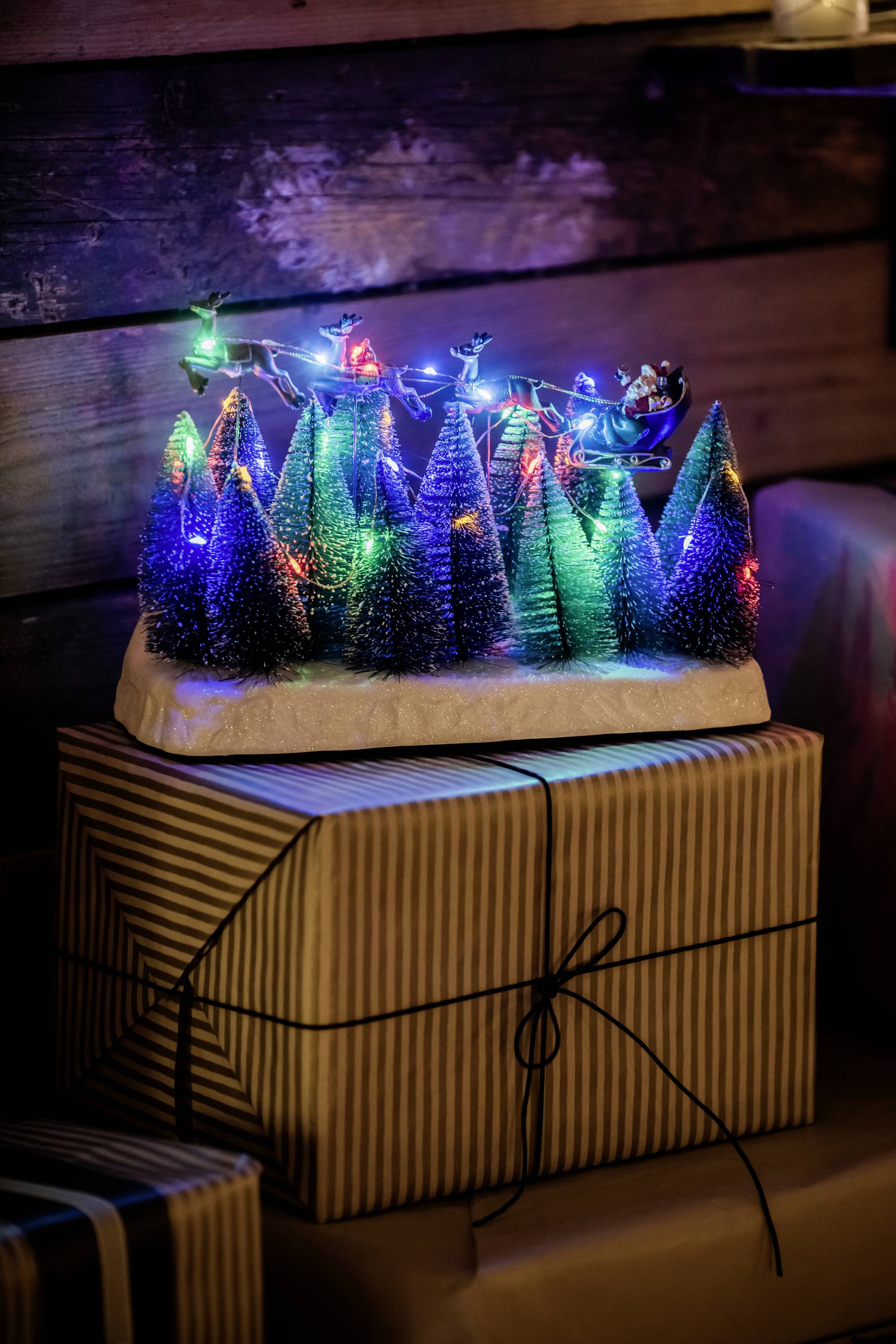 Bunte LED-Lichterketten schmücken kleine Tannenbäume auf einem Geschenk, umgeben von weiteren Paketen. Festliche Weihnachtsdekoration.