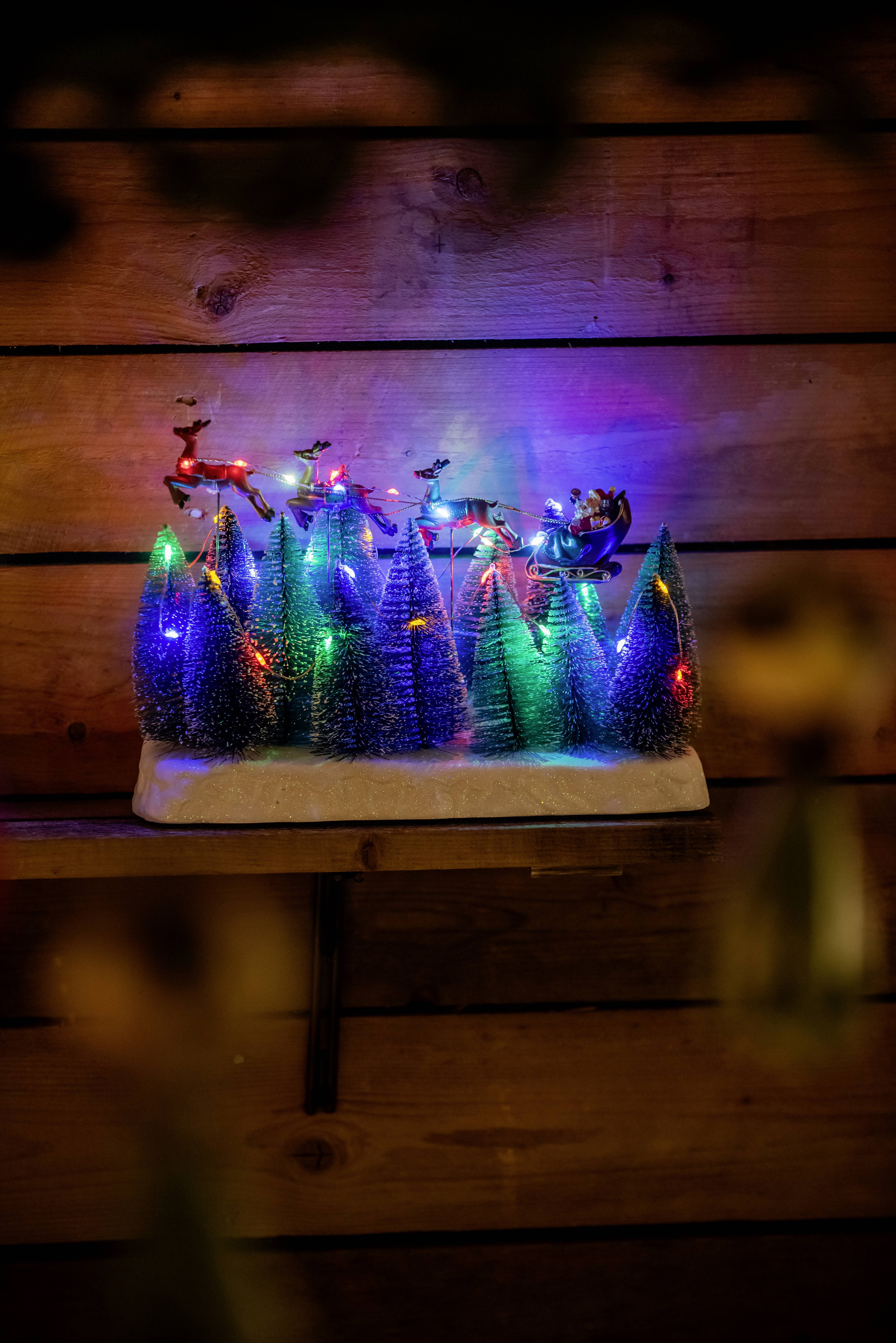 Kleine, leuchtende Weihnachtsbaum-Dekorationen mit bunten Lichtern. Rentierschlitten oben. Holzrückwand im Hintergrund.