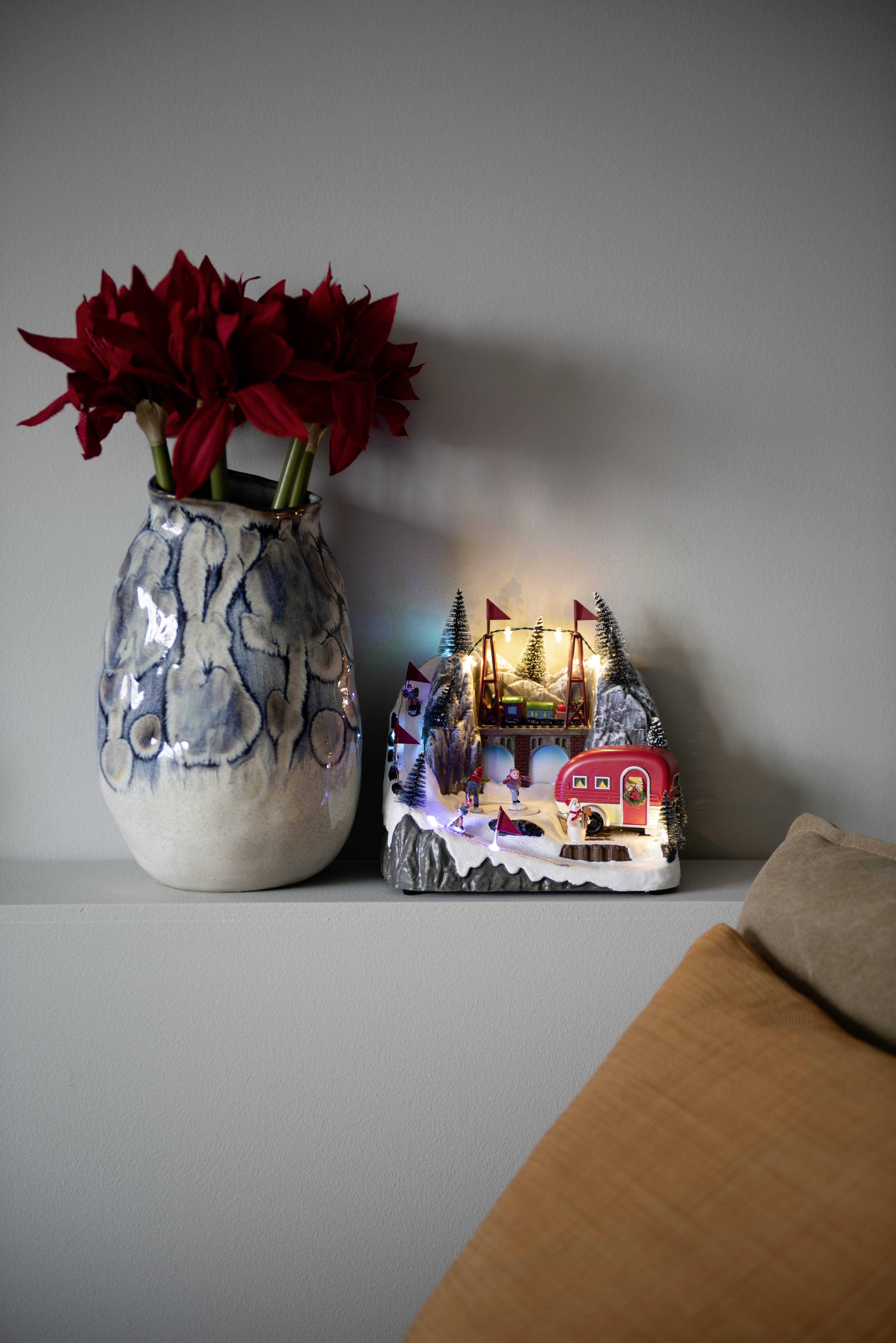 Vase mit roten Blumen und Miniaturdorf mit Beleuchtung auf einem Regal, daneben ein beiges Kissen. Gemütliche Wohnatmosphäre.