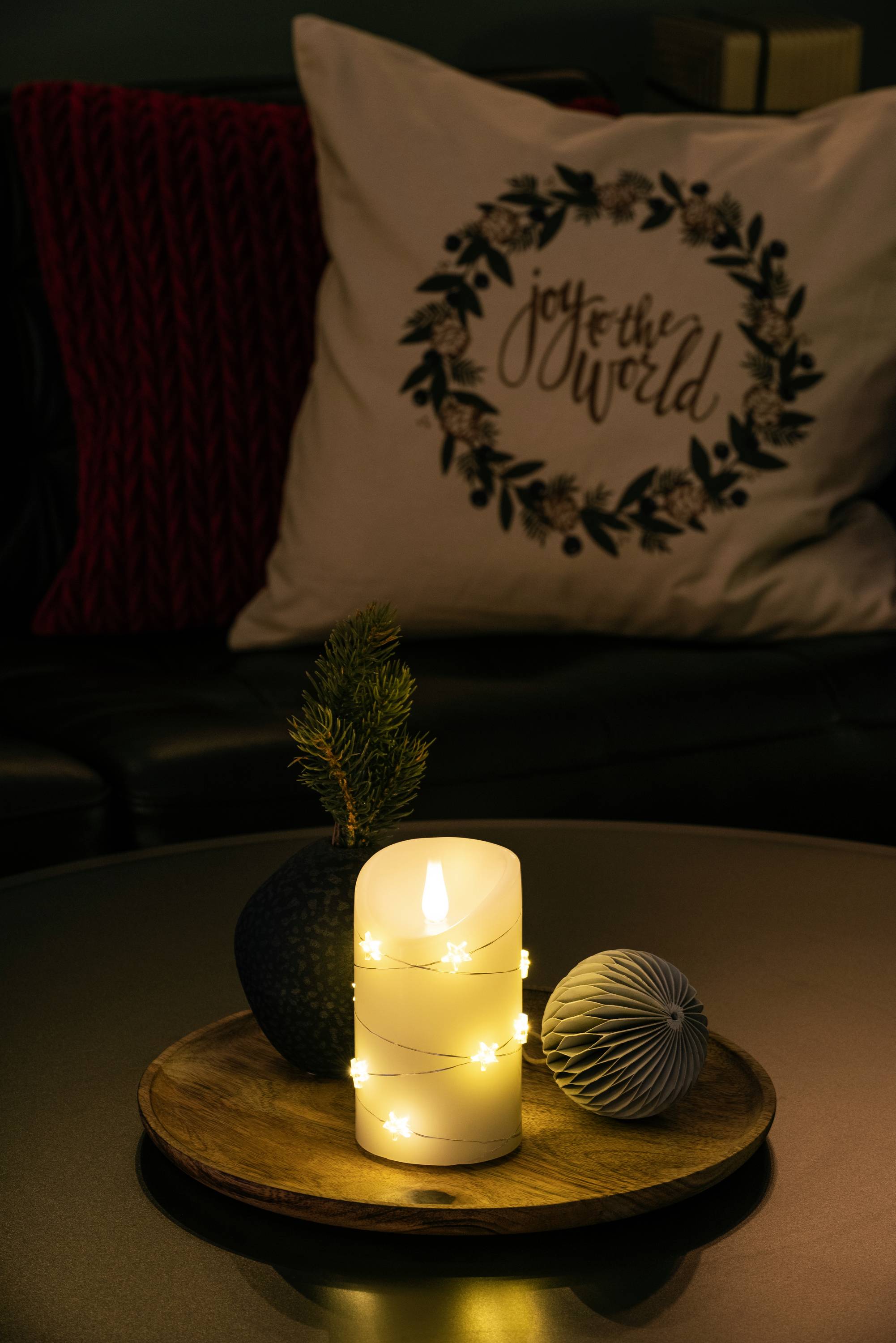 Eine brennende Kerze mit Sternenlichtern auf einem Holztablett neben Dekorationen. Im Hintergrund ein Kissen mit 'joy to the world'.