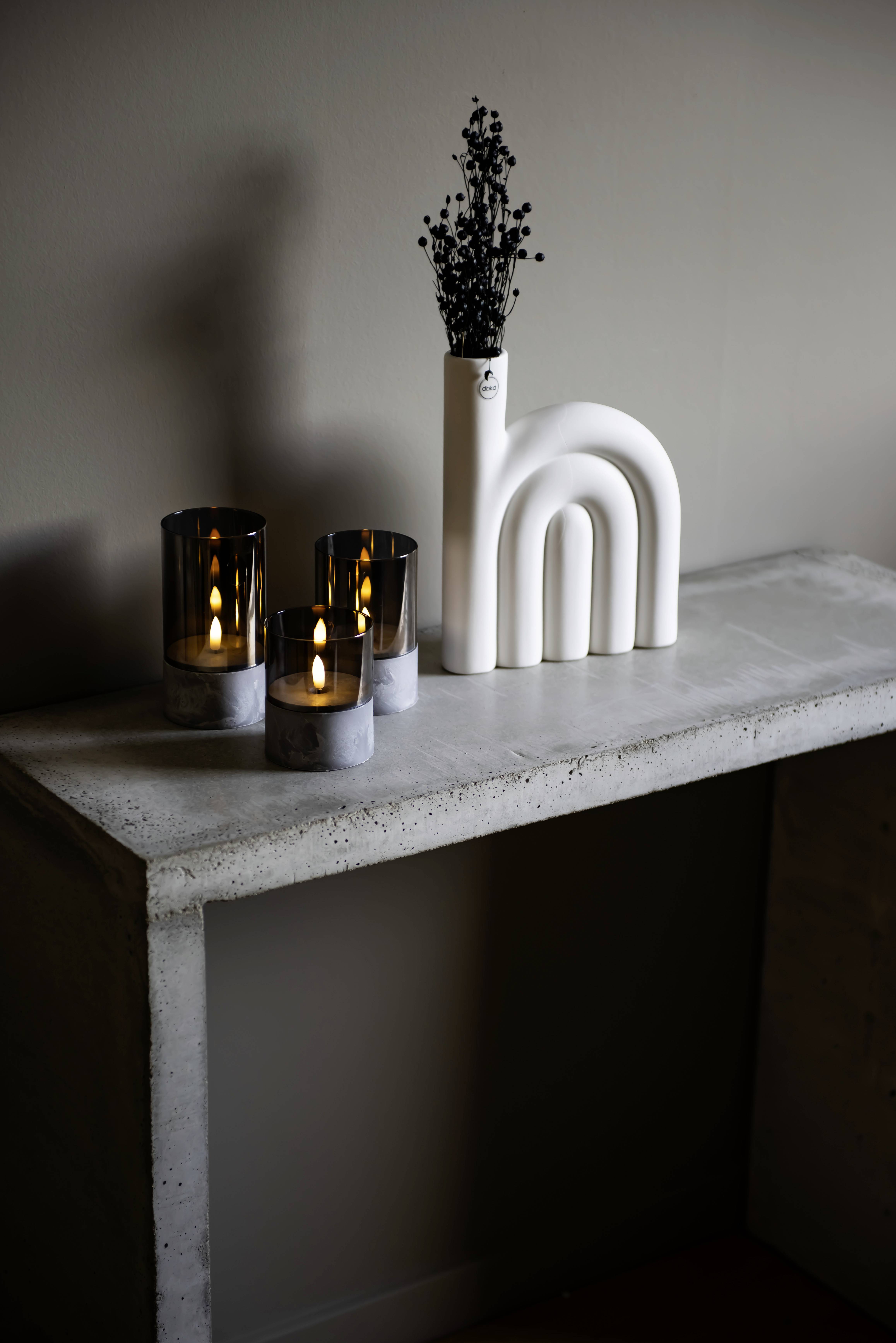 Eine minimalistische Betonkonsolentisch mit drei brennenden Kerzen in modernen Glasgefäßen und einer weißen Vase mit getrockneten Blumen.